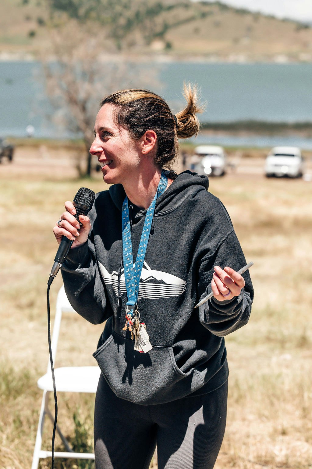 A woman speaks into a microphone outdoors, wearing a hoodie and lanyard, with a scenic lake background and parked cars.