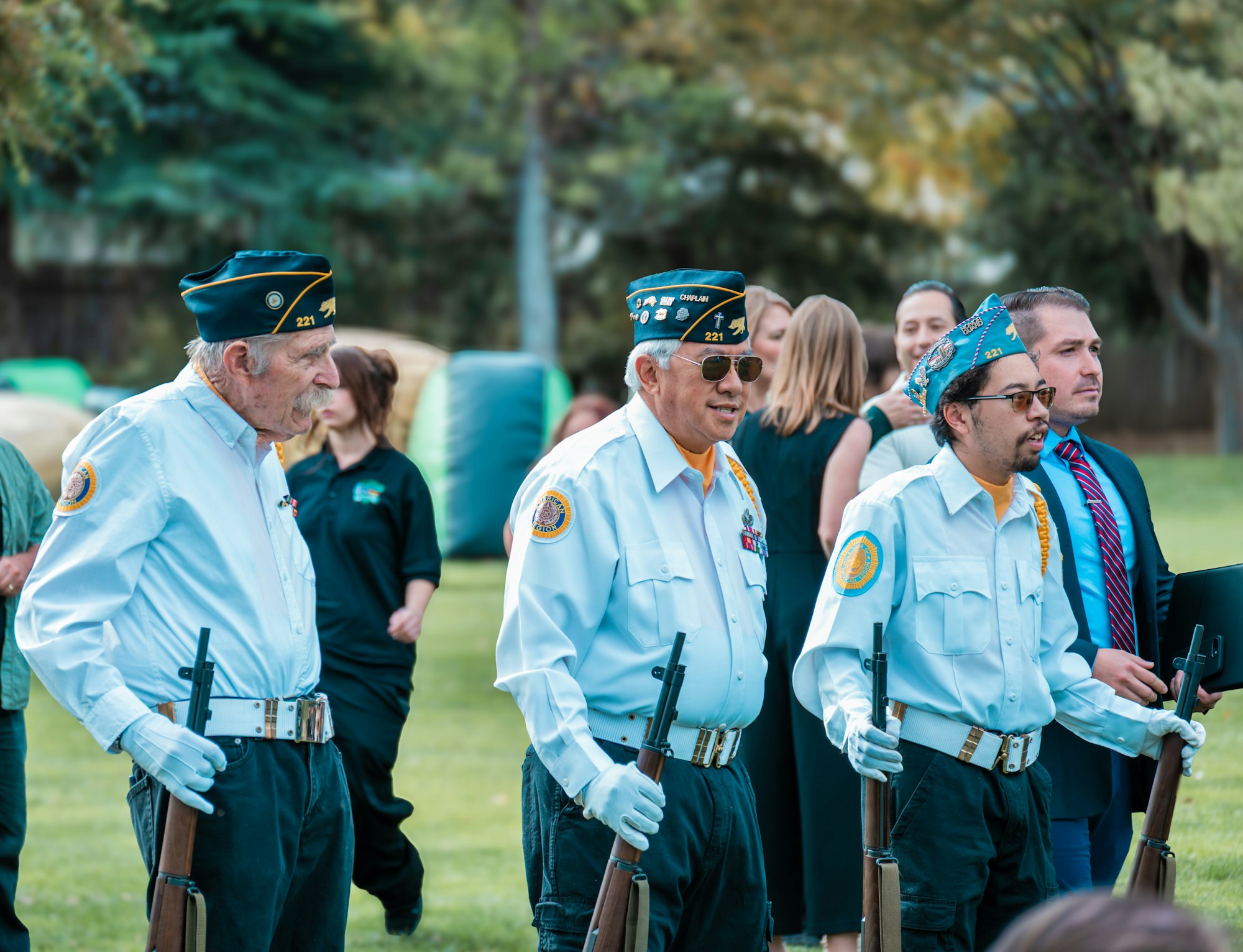 Veterans in uniform holding rifles stand in formation at a public event, surrounded by other attendees.
