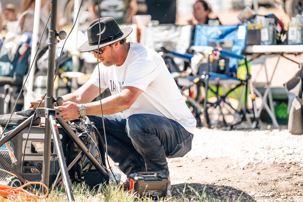Man in a hat working on outdoor film set equipment, with chairs and gear in background.