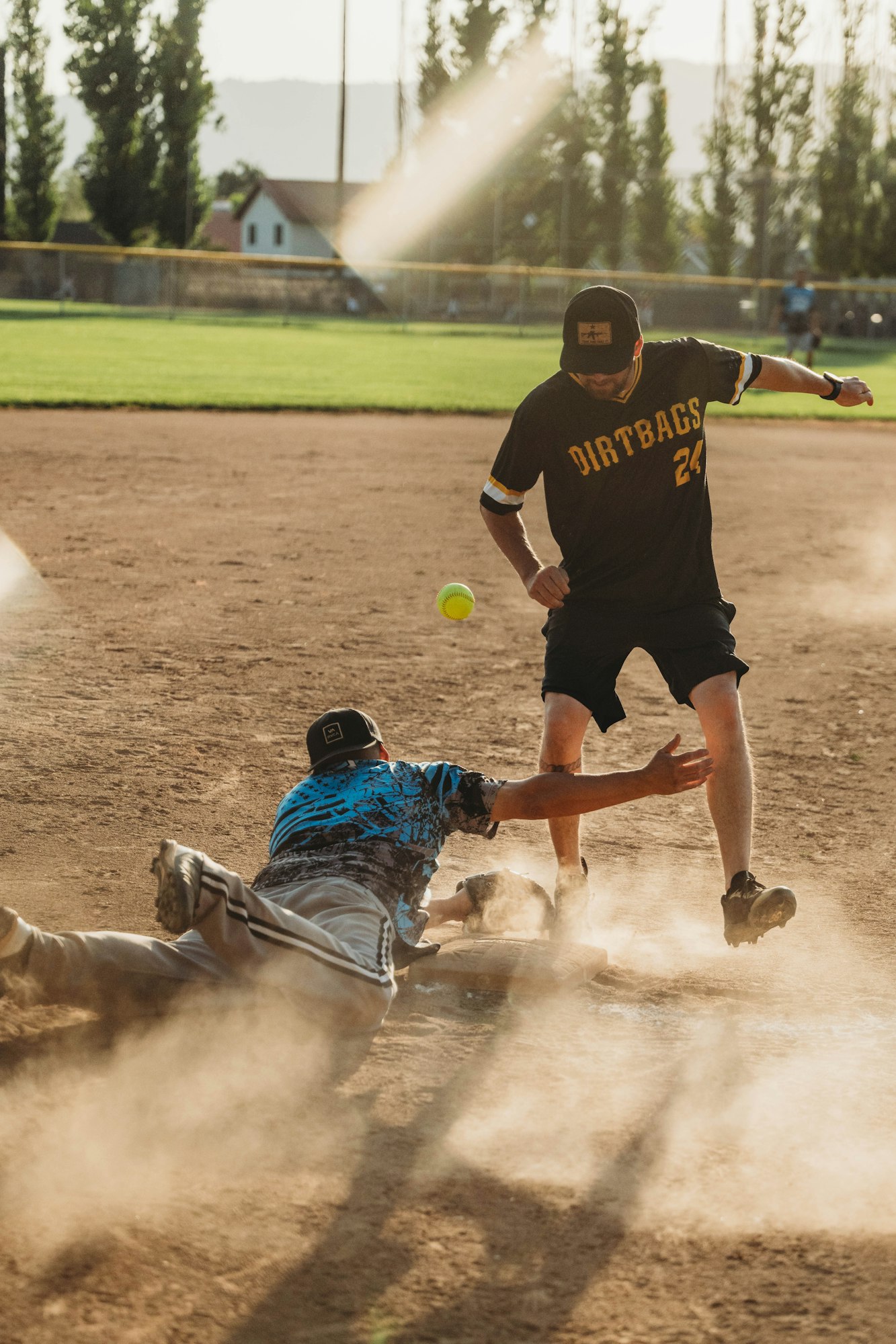 A baseball player slides into base while another prepares to catch the ball, dust billowing, during a sunny game.