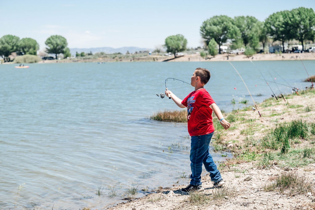 A boy fishing by a lake, rods lined up on shore, trees in background, and a kayak in the distance.
