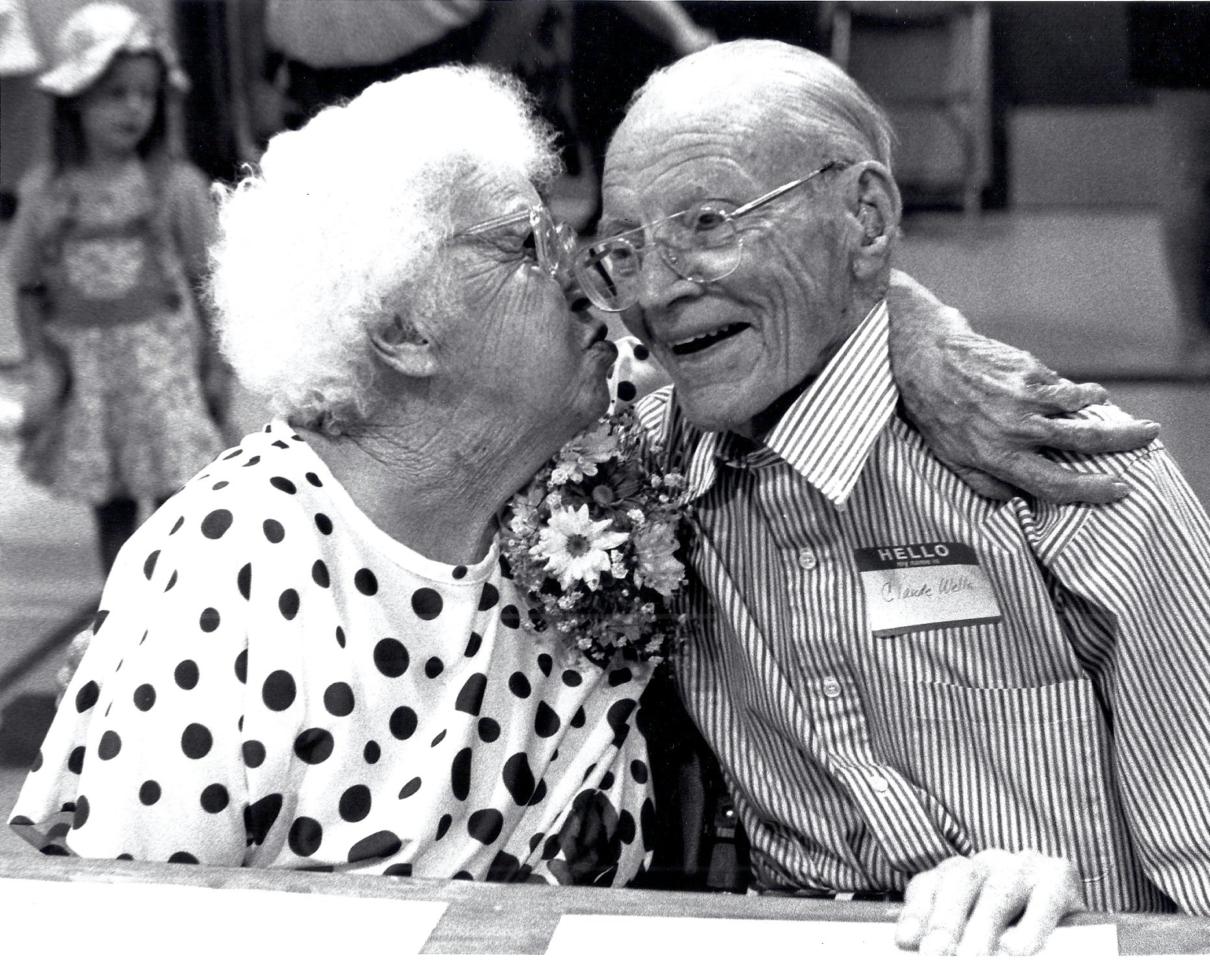 An elderly couple shares an affectionate moment; the woman kisses the man's cheek while embracing him.