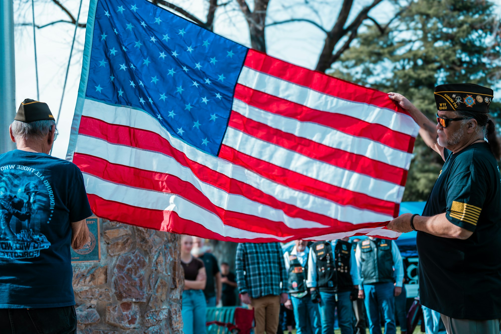 Two people holding an American flag during a gathering, with onlookers in the background.
