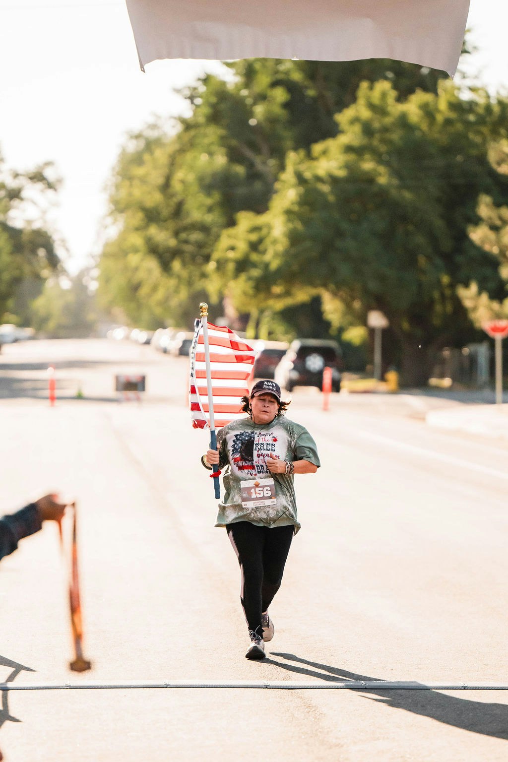 Person running with a U.S. flag, wearing number 156, on an empty street under greenery.