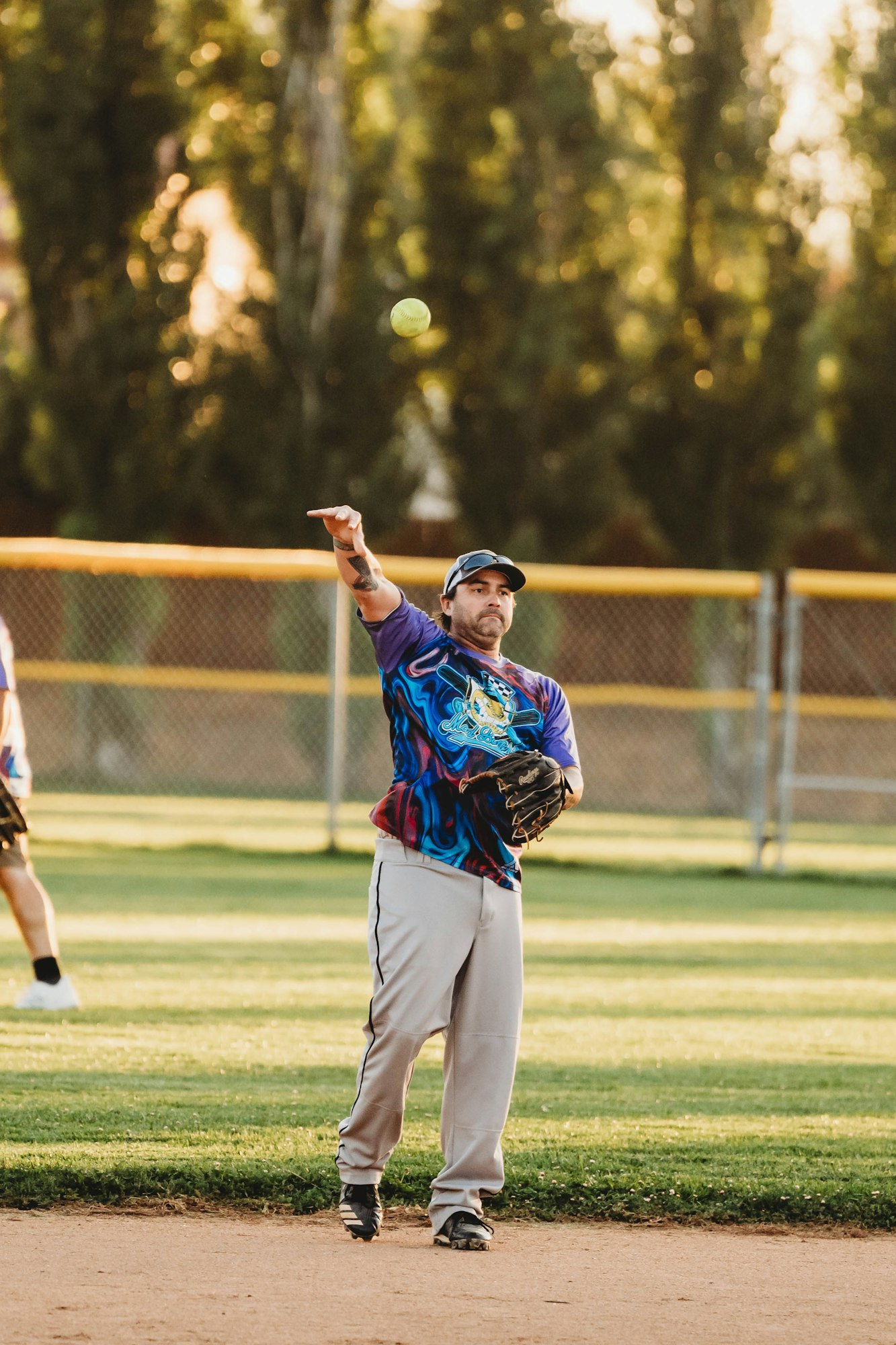 A baseball player throwing a ball, standing on a dusty field, with trees in the background.