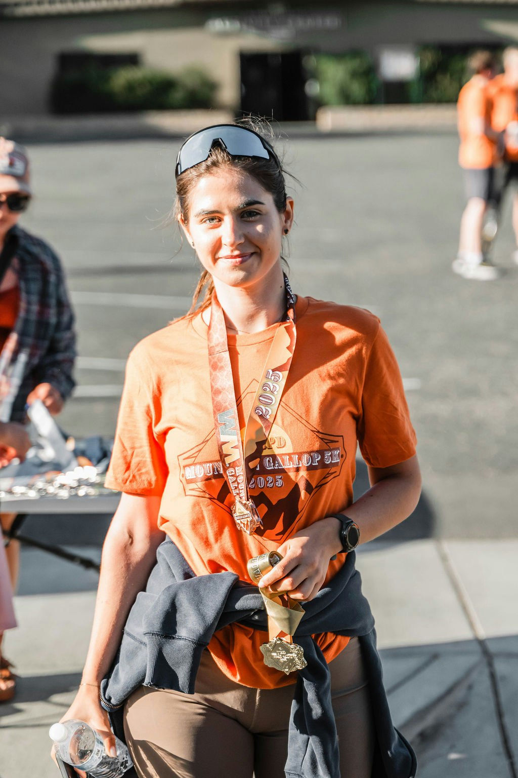 Person in orange shirt with medal and sunglasses, holding a water bottle at an outdoor event or race.