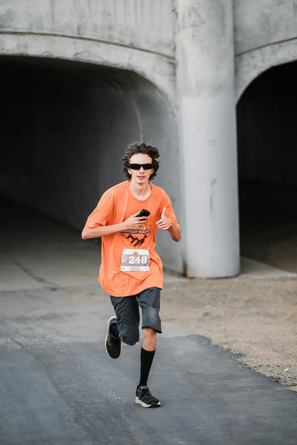 A person running outdoors with sunglasses, wearing an orange shirt, a race bib number “248,” giving a thumbs-up gesture.