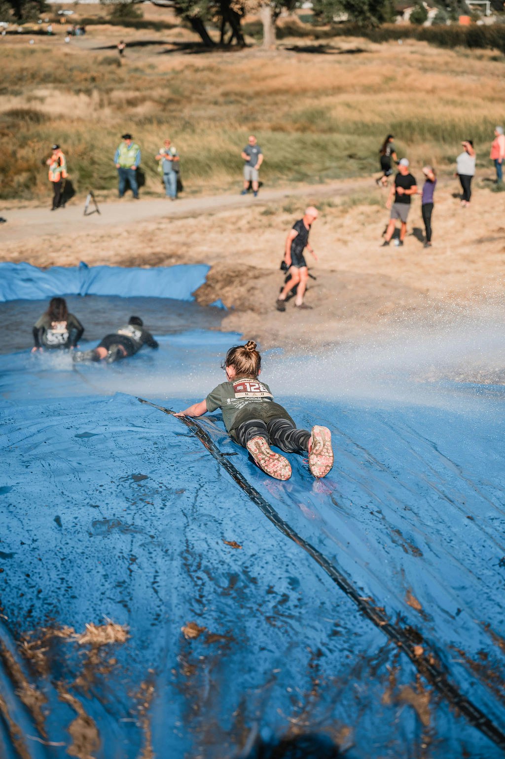 A person slides down a wet, blue tarp, while others wait at the bottom. Outdoor setting with spectators around.