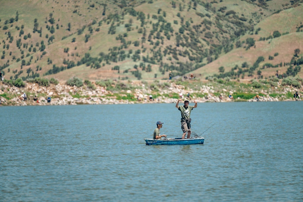 Two people on a small boat fishing in a lake with hilly background.