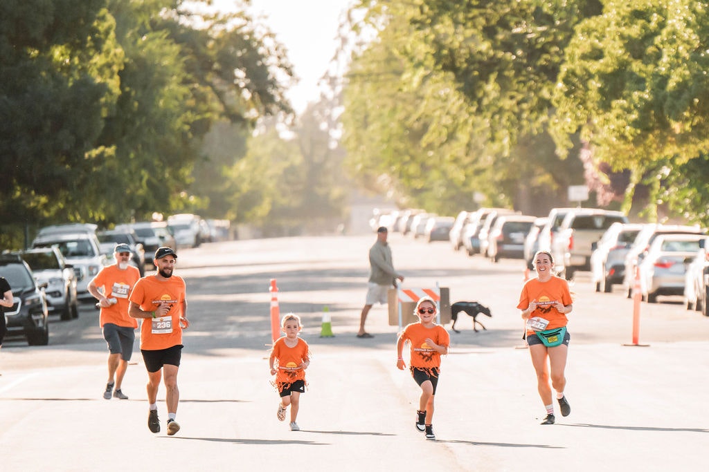 A group of people running on a street wearing matching orange shirts, with cars parked alongside and trees in the background.