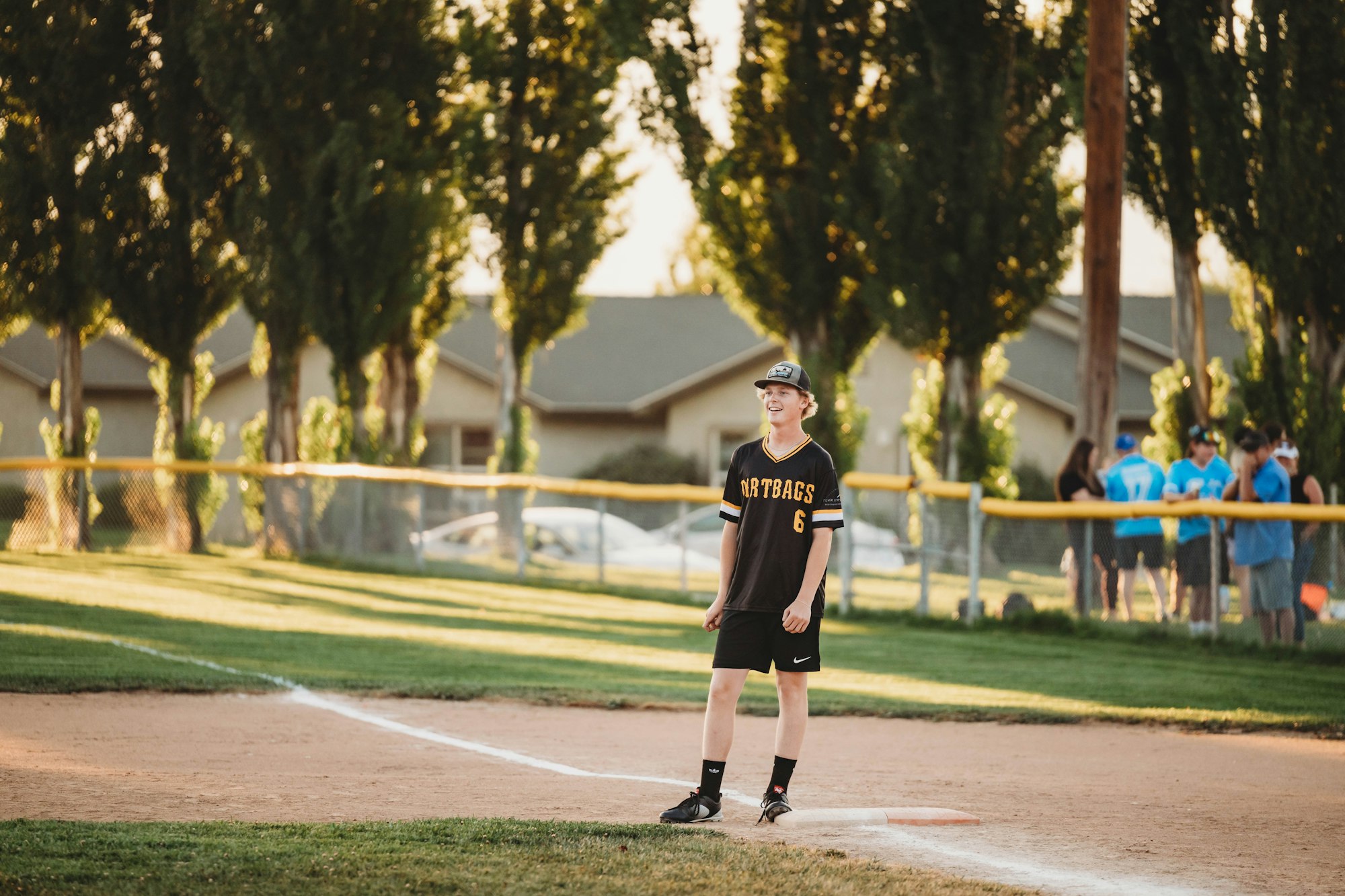 A person in a baseball uniform stands on a field with trees and spectators in the background.