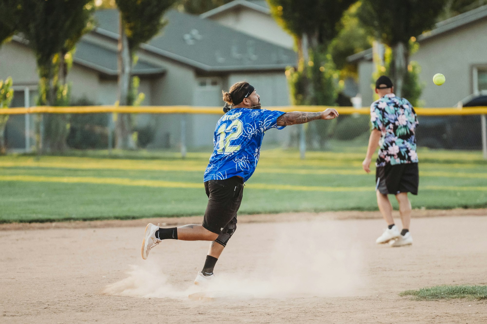 A player in a blue jersey throws a ball on a dusty field, while another person in a floral shirt stands nearby.