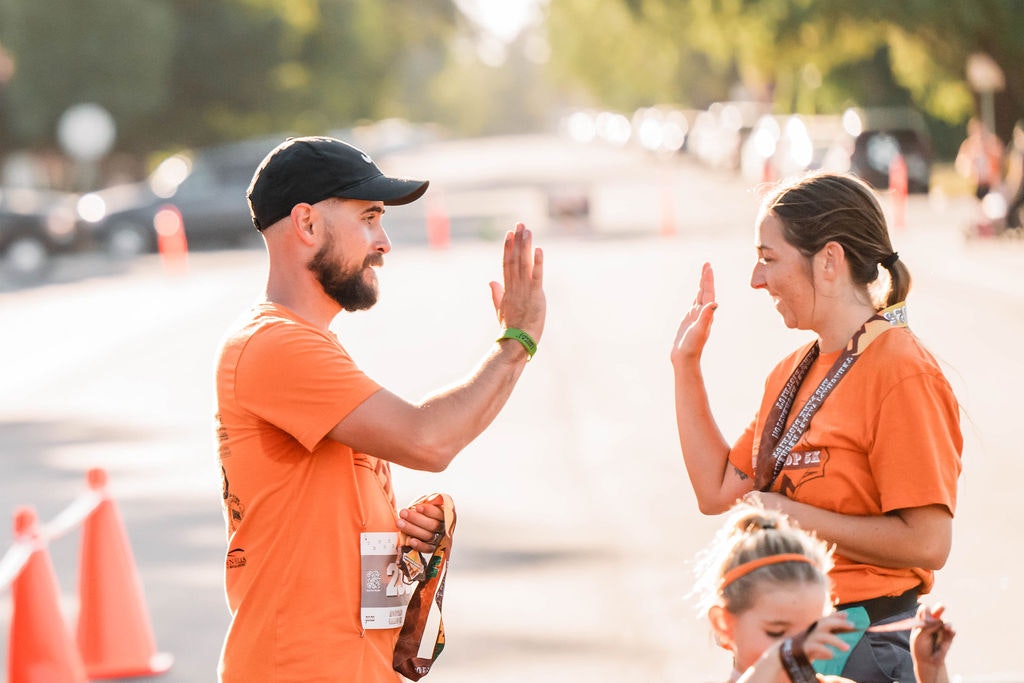 Two people in orange shirts high-five each other outdoors, smiling. A child stands in the foreground.