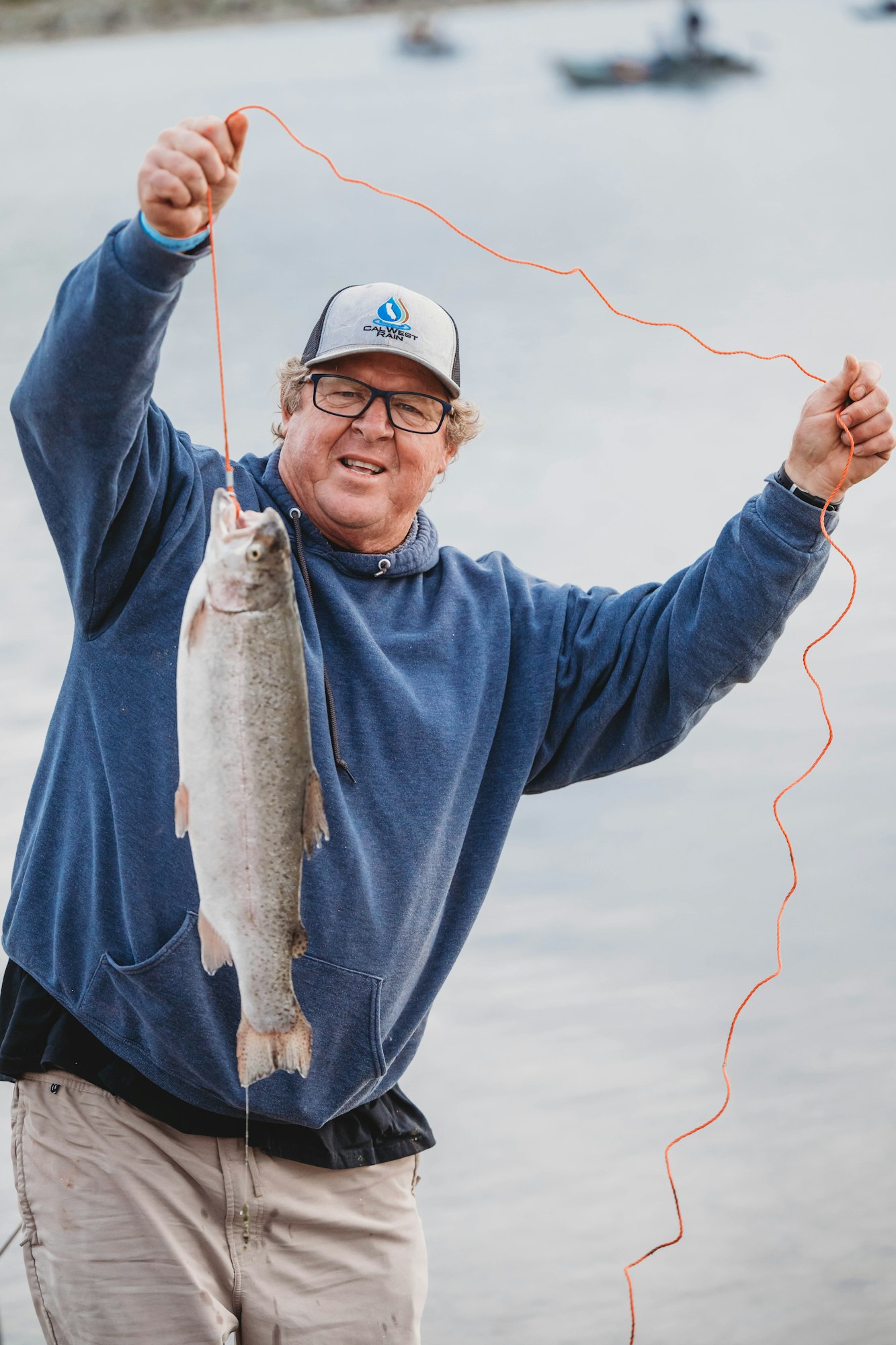Person holding a fish with a string by a body of water, wearing a blue hoodie and cap.