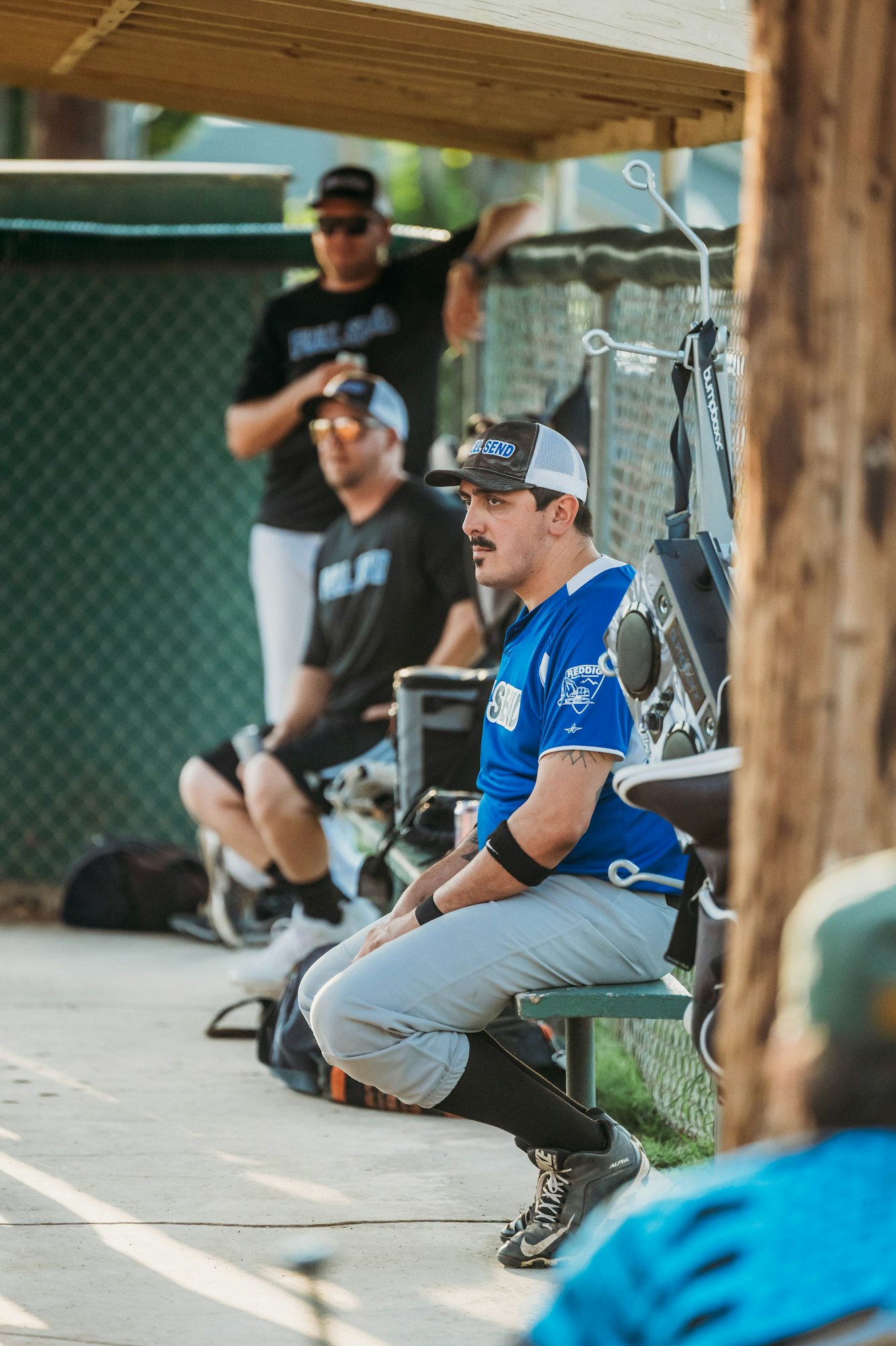 Baseball players in dugout, one squatting in foreground, focused, wearing blue jersey and cap.