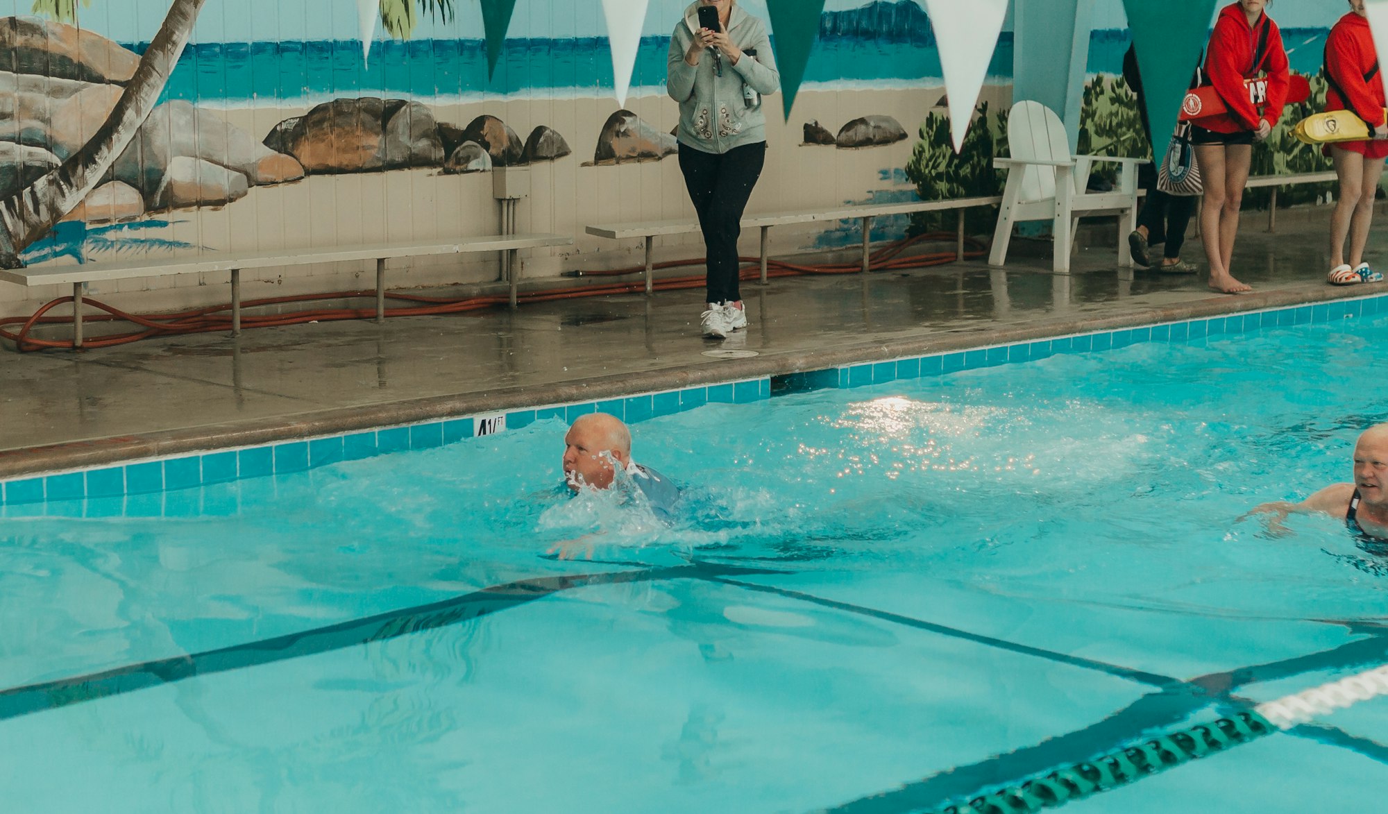 Indoor pool with swimmers, a person taking a photo, lifeguards, and a tropical mural backdrop.