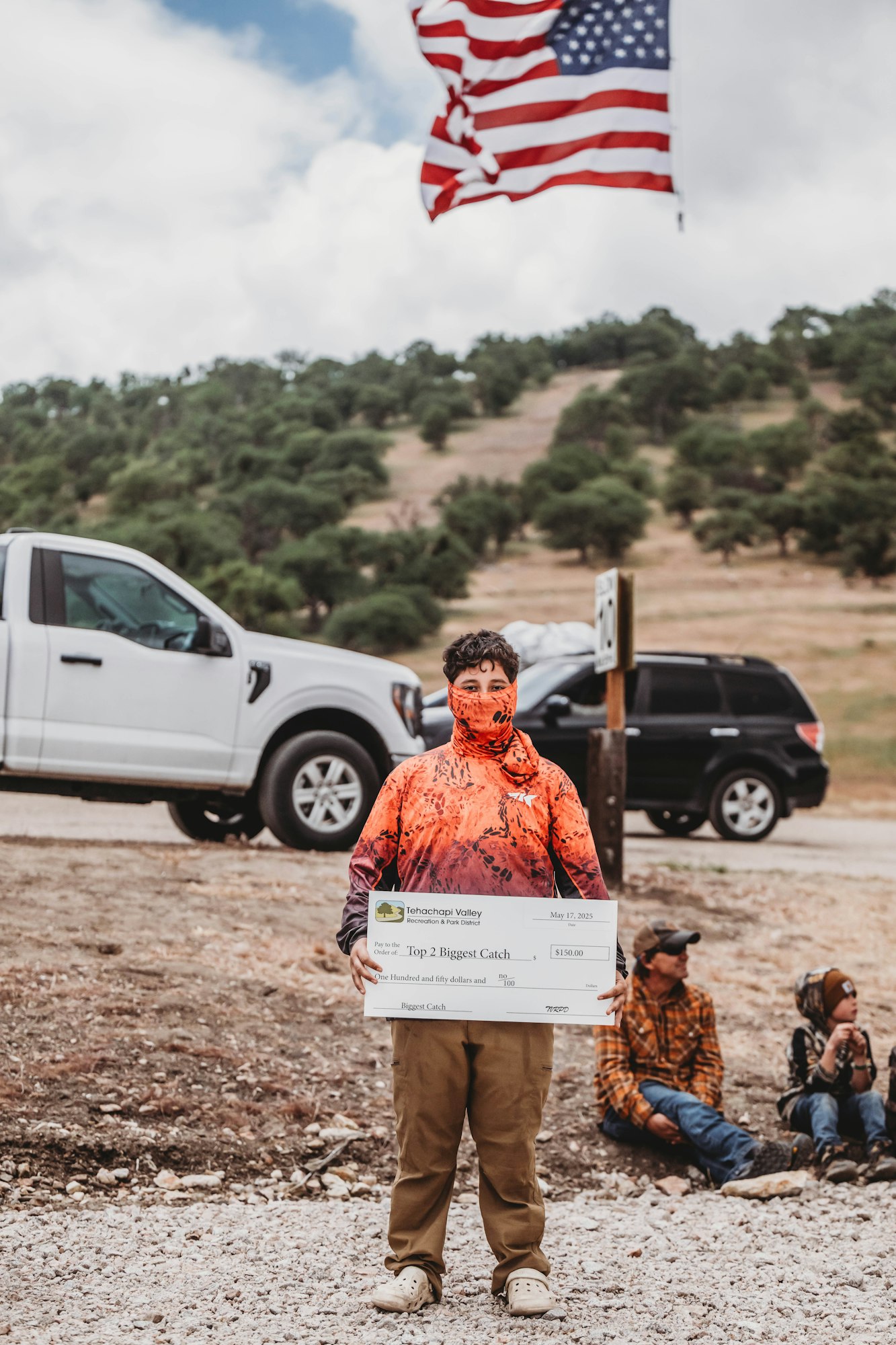A person in an orange outfit holds a large check for a fishing contest prize. An American flag waves in the background.