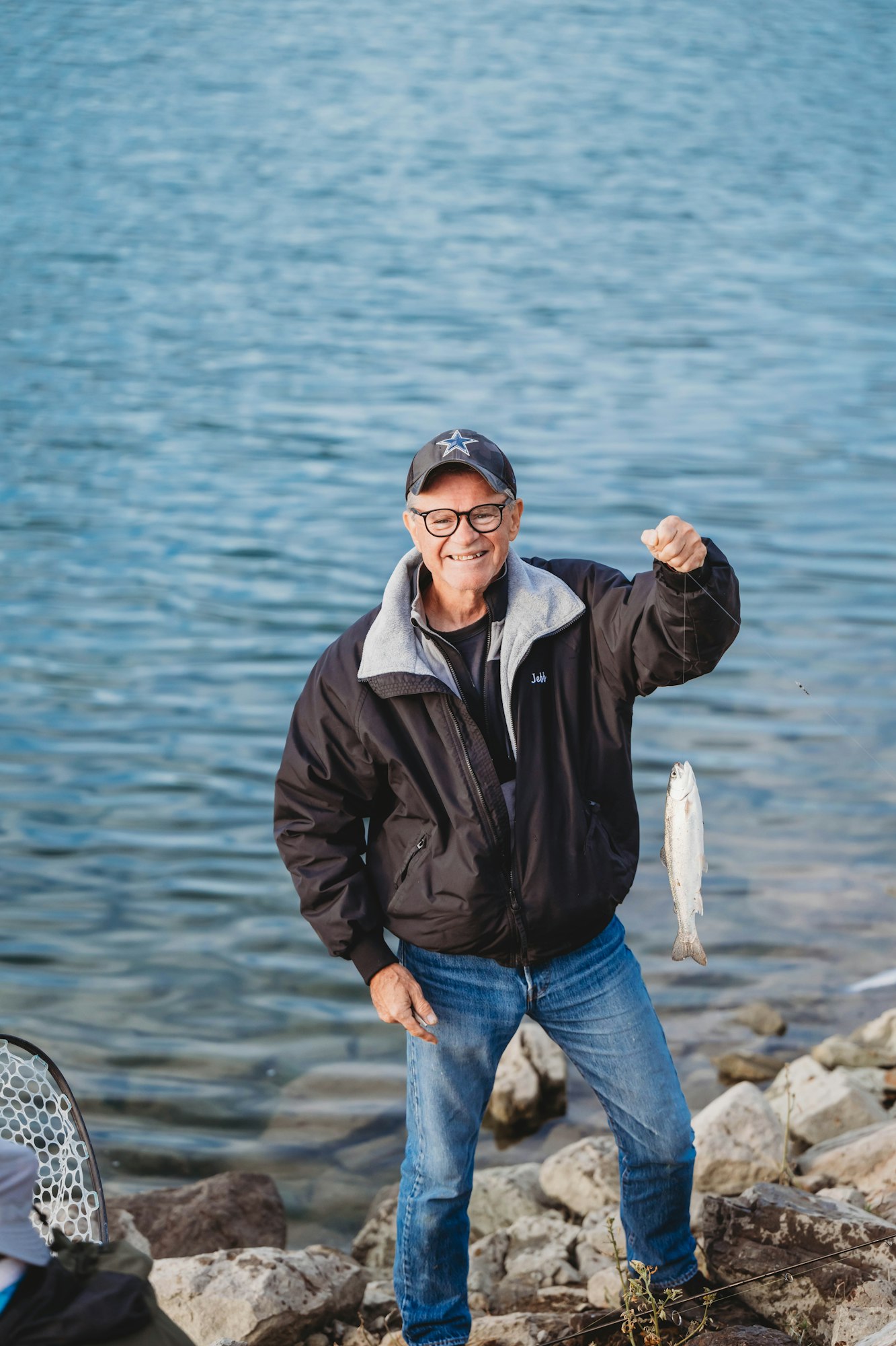 A person by a lake holding a fish, smiling, wearing a cap and jacket.