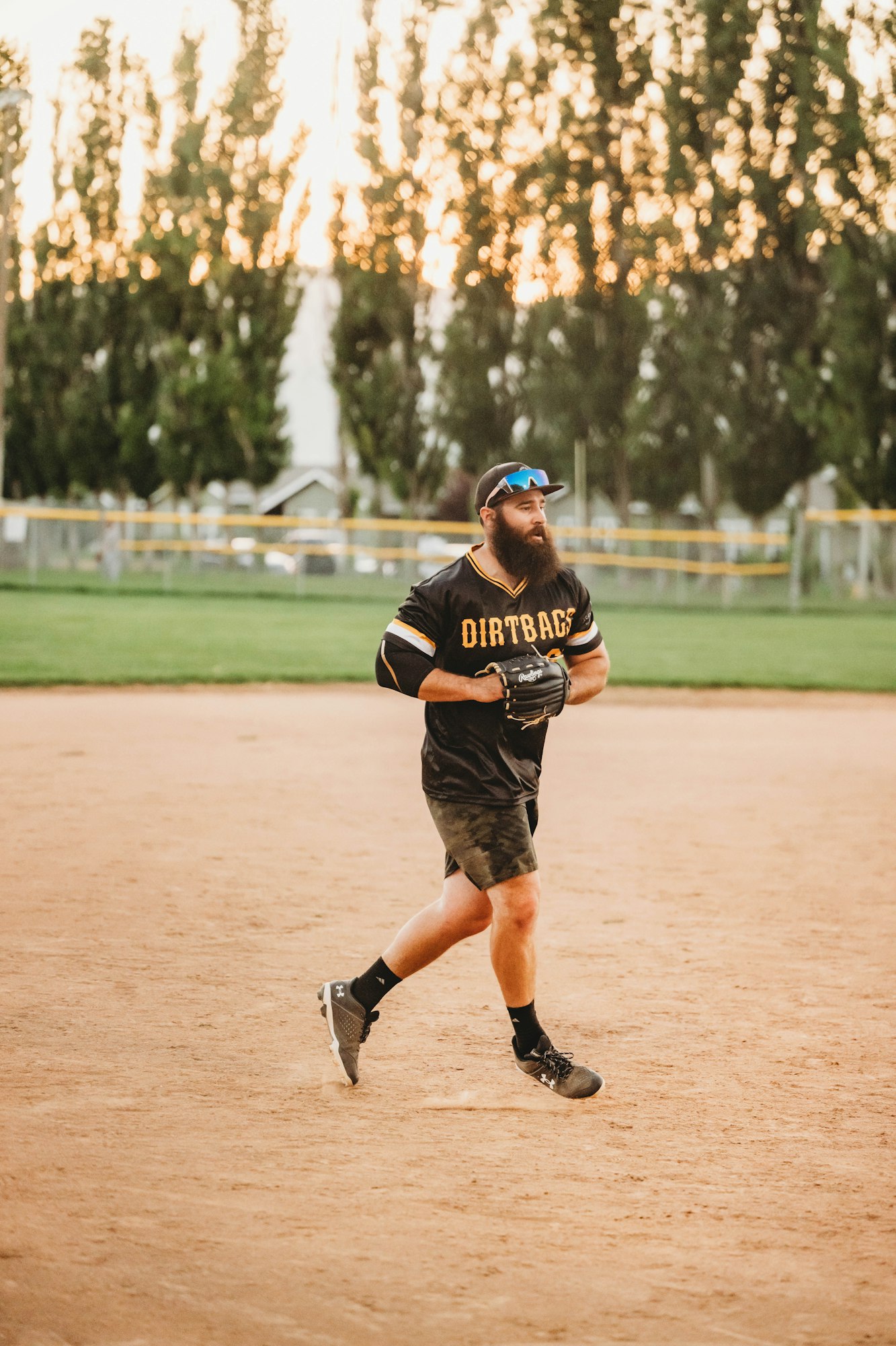 A bearded softball player running on a dirt field with trees and sunset in the background.