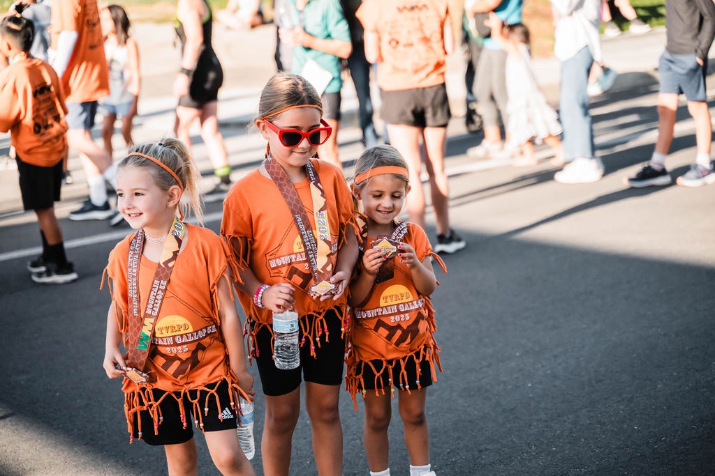 Three kids in orange shirts and medals standing together, with a crowd in the background.