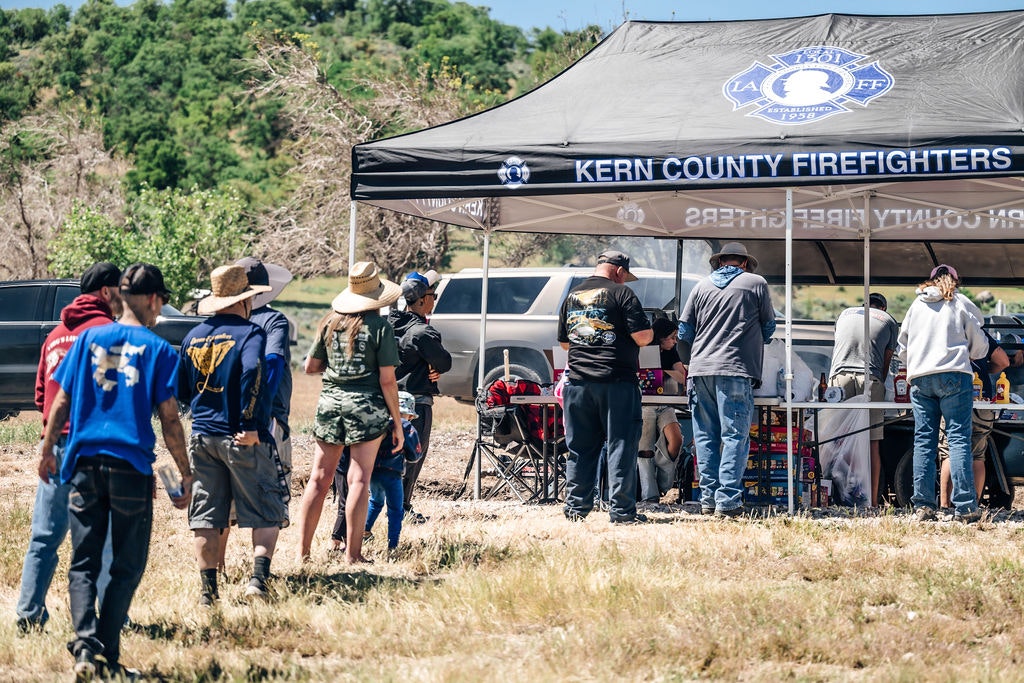 People gathered around a tent with "Kern County Firefighters" banner, likely at an outdoor event.