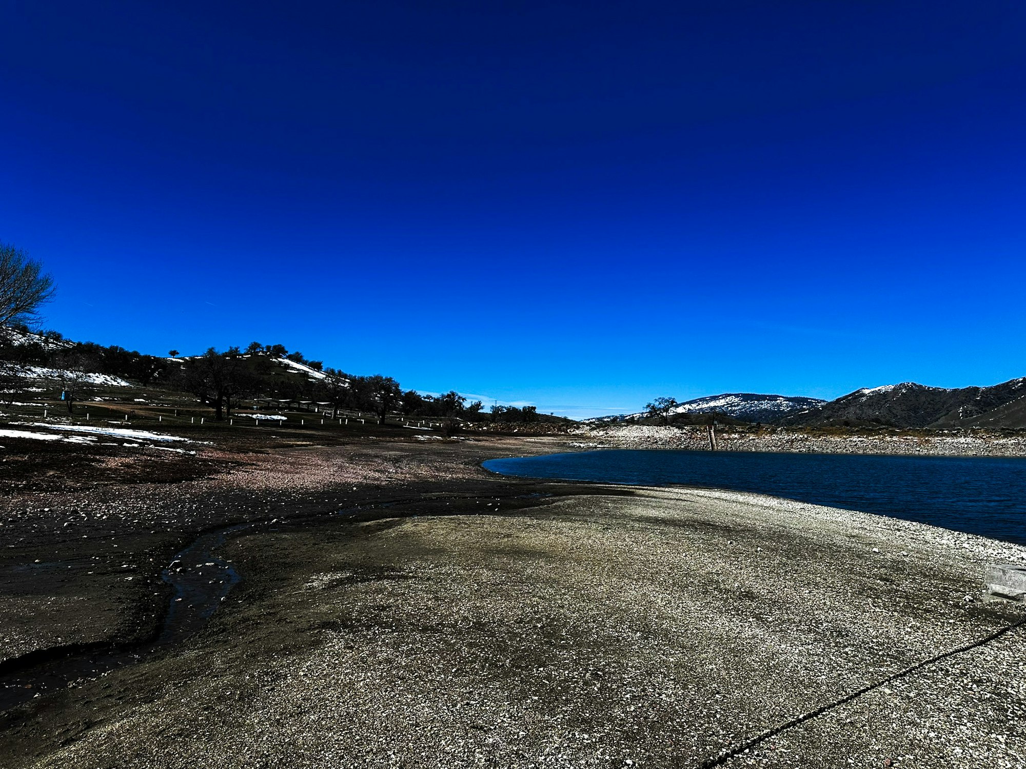 A lakeshore with a gravel path, water, snow-dappled hills, and a clear blue sky.