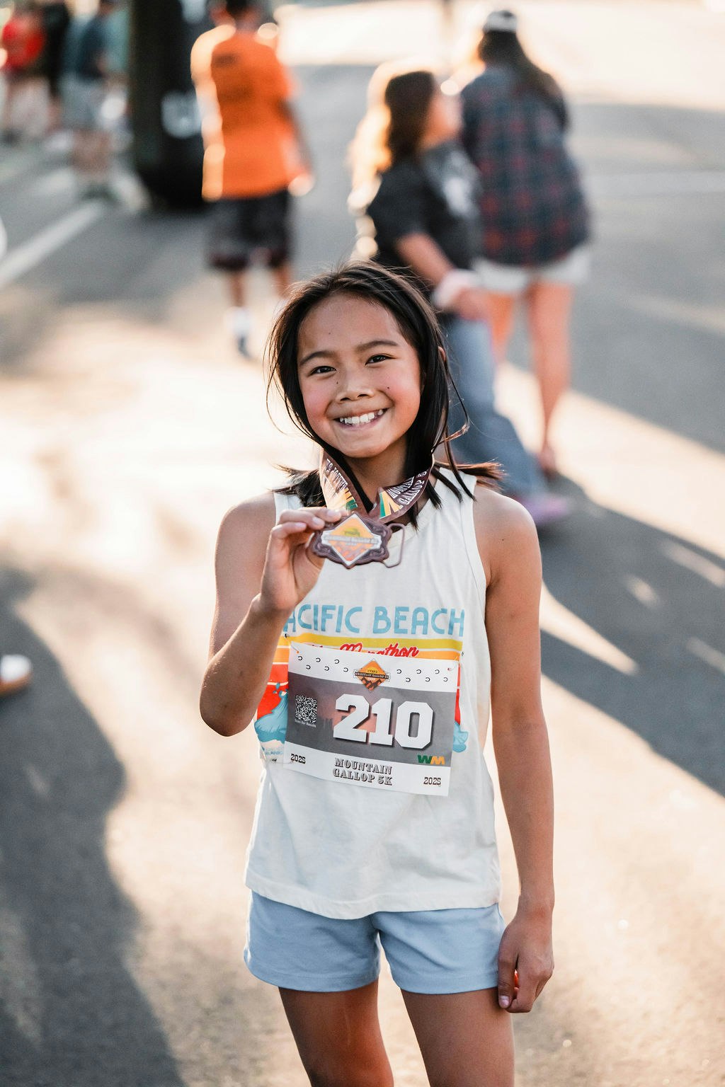 A young girl smiling, wearing a marathon bib number 210, holding a medal.