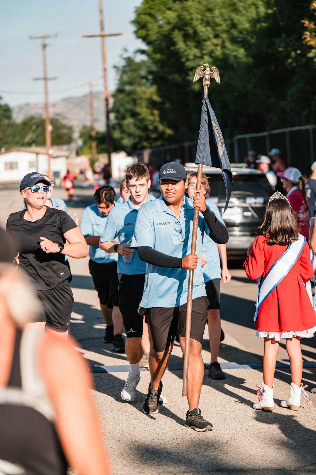 People march in a parade, wearing blue shirts, one holds a flag. A girl in a red dress and sash watches.