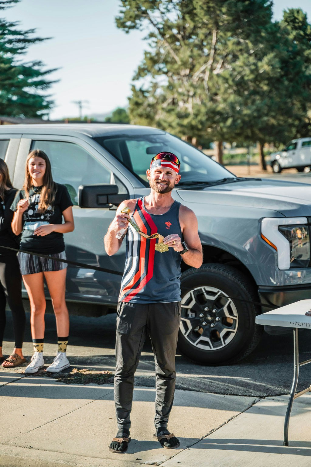 A person is standing in front of a vehicle holding two medals, others are in the background, outdoors.