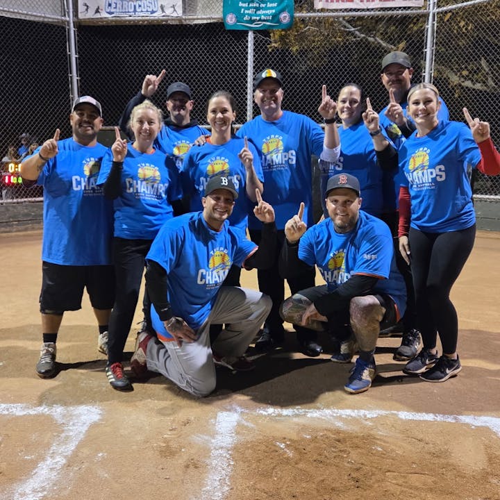 A group of people wearing blue "Champs" shirts, posing on a baseball field, showing number one gesture.