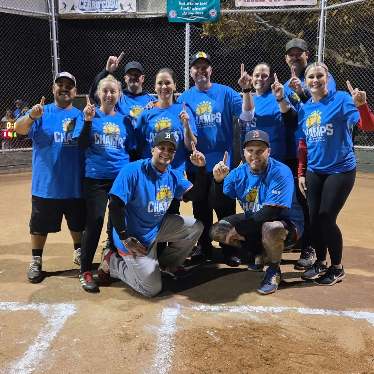 A group of people wearing blue "Champs" shirts, posing on a baseball field, showing number one gesture.