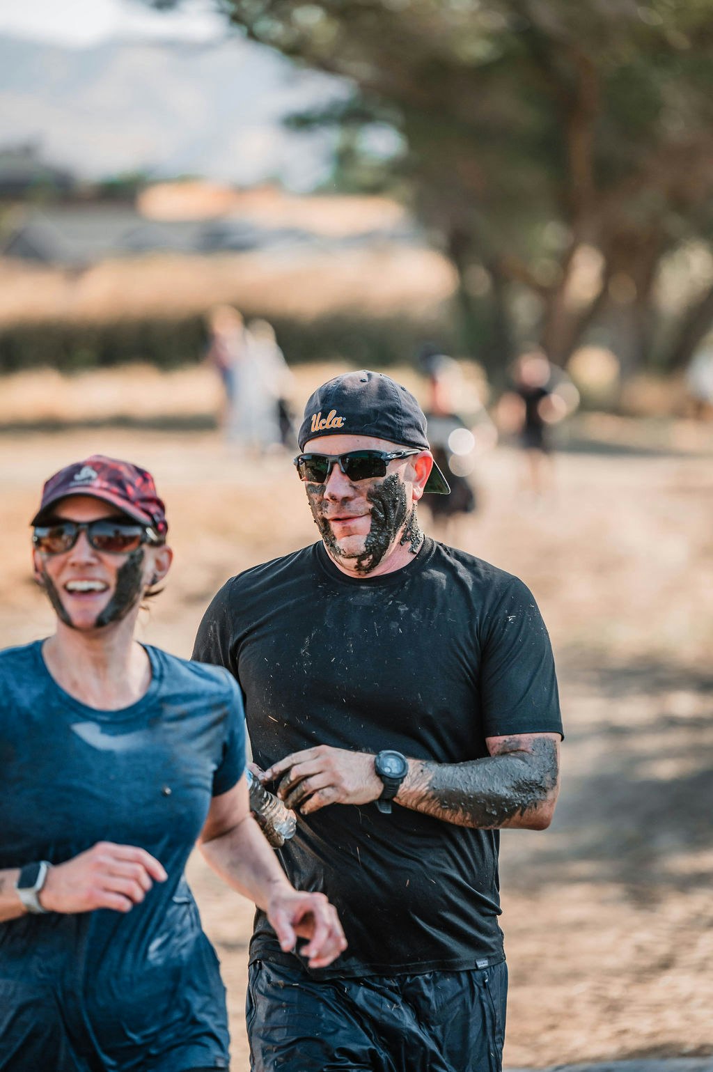 Two people running outdoors with mud on their faces, wearing sunglasses and sporty attire.