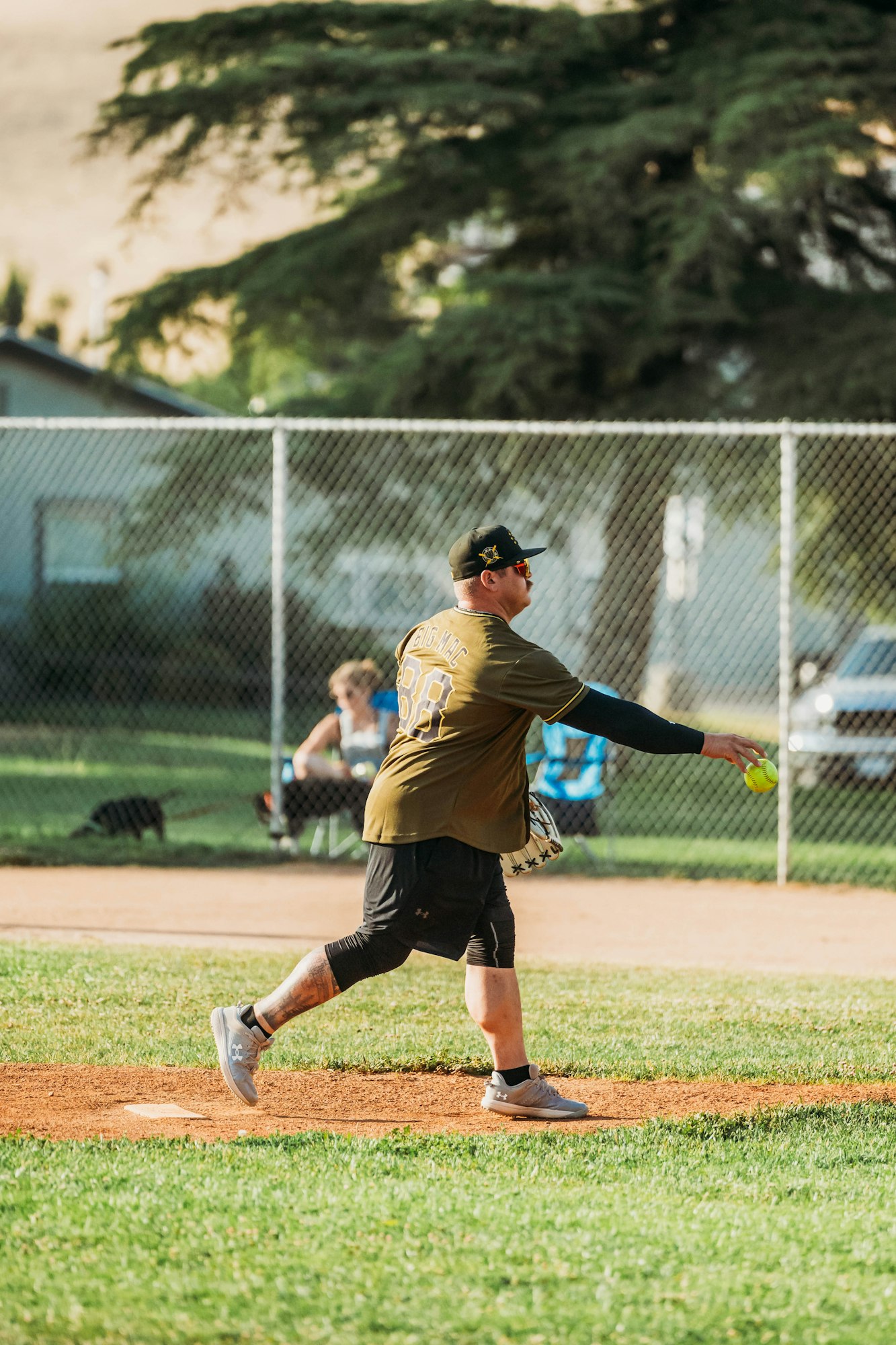 A person is pitching a softball in a park with trees and a fence in the background.