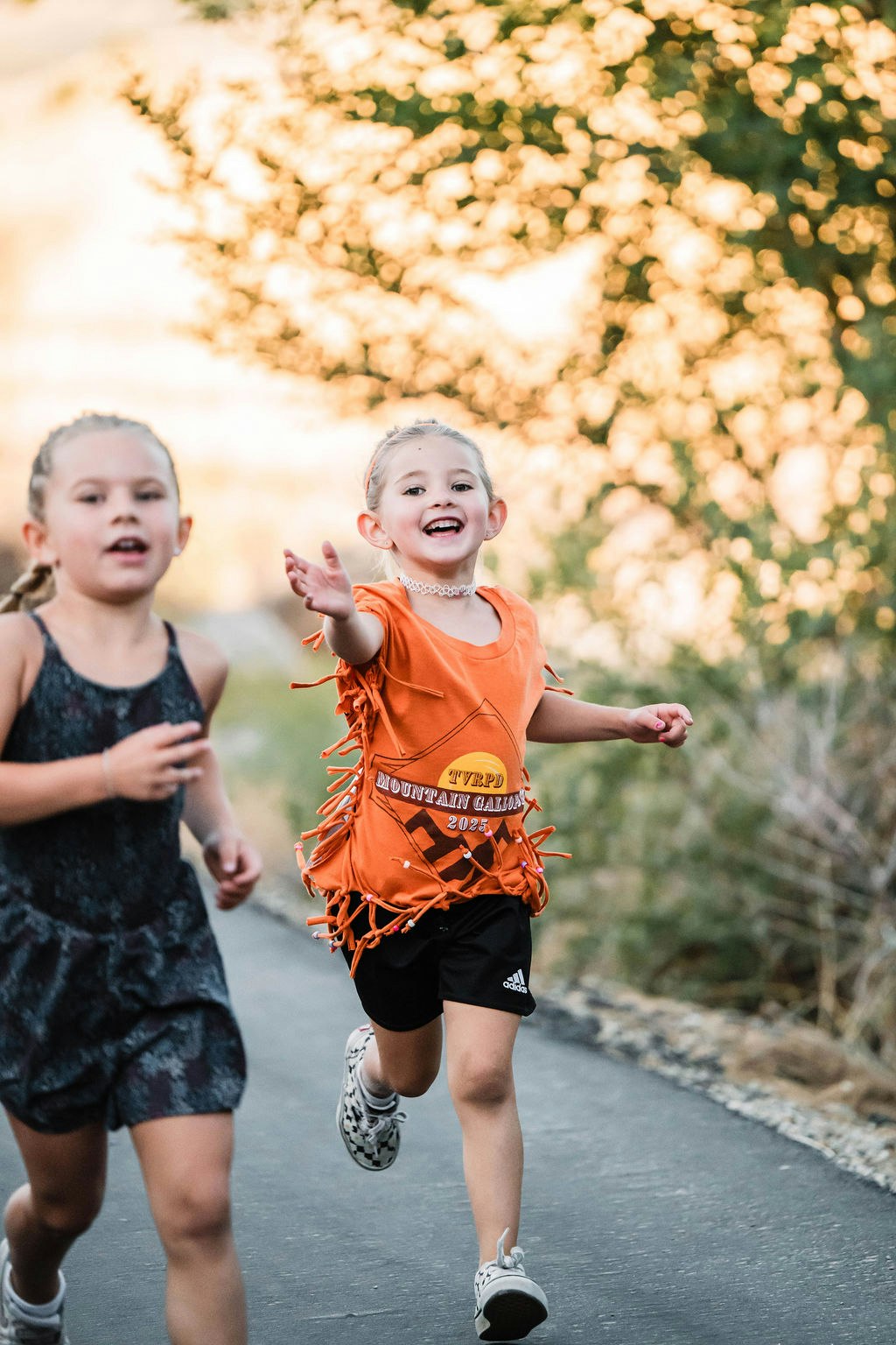 Two children running outdoors, one wearing an orange shirt and smiling, with trees in the background.