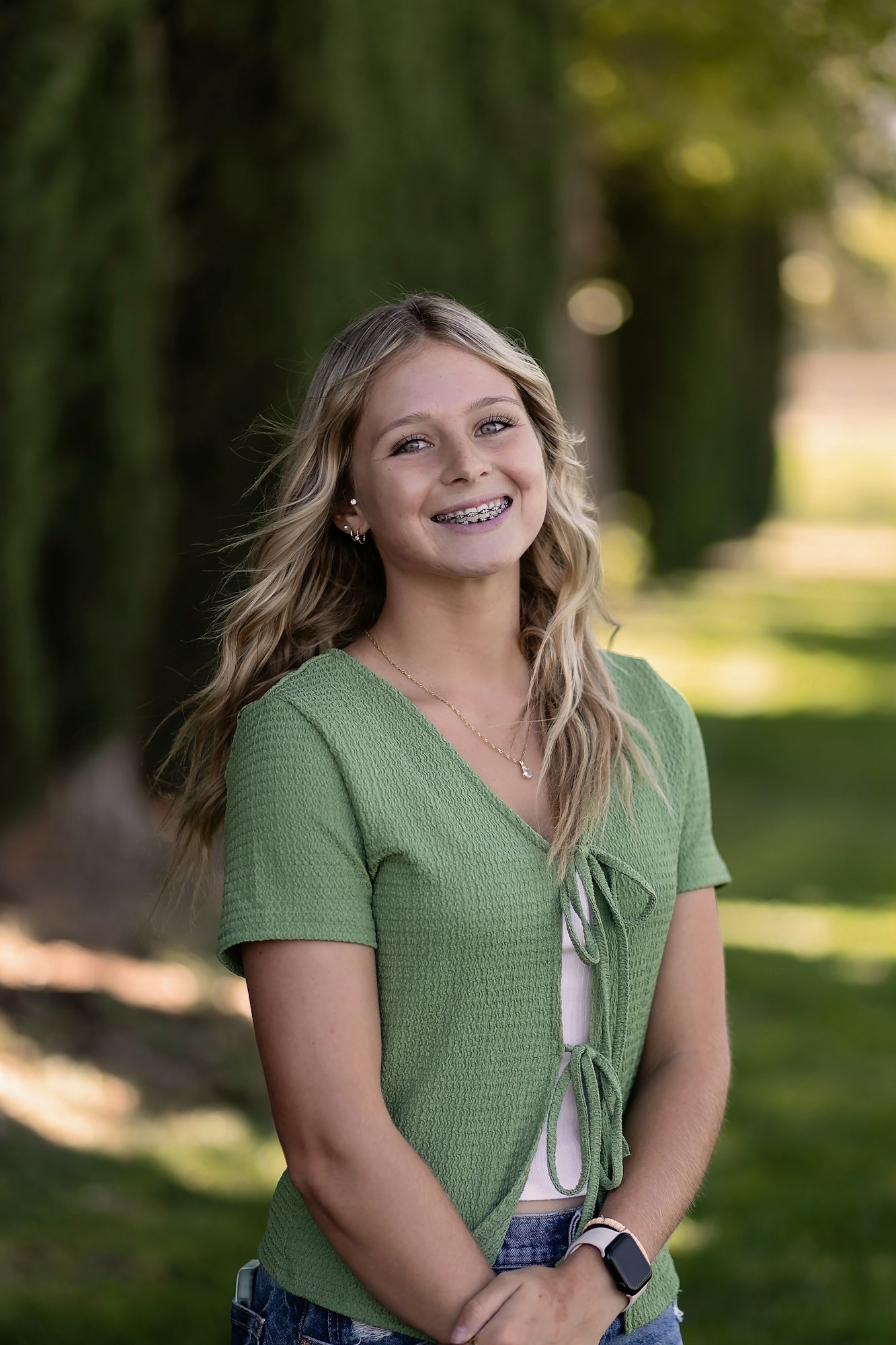 A person smiling outdoors, wearing a green top and braces, with trees and grass in the background.
