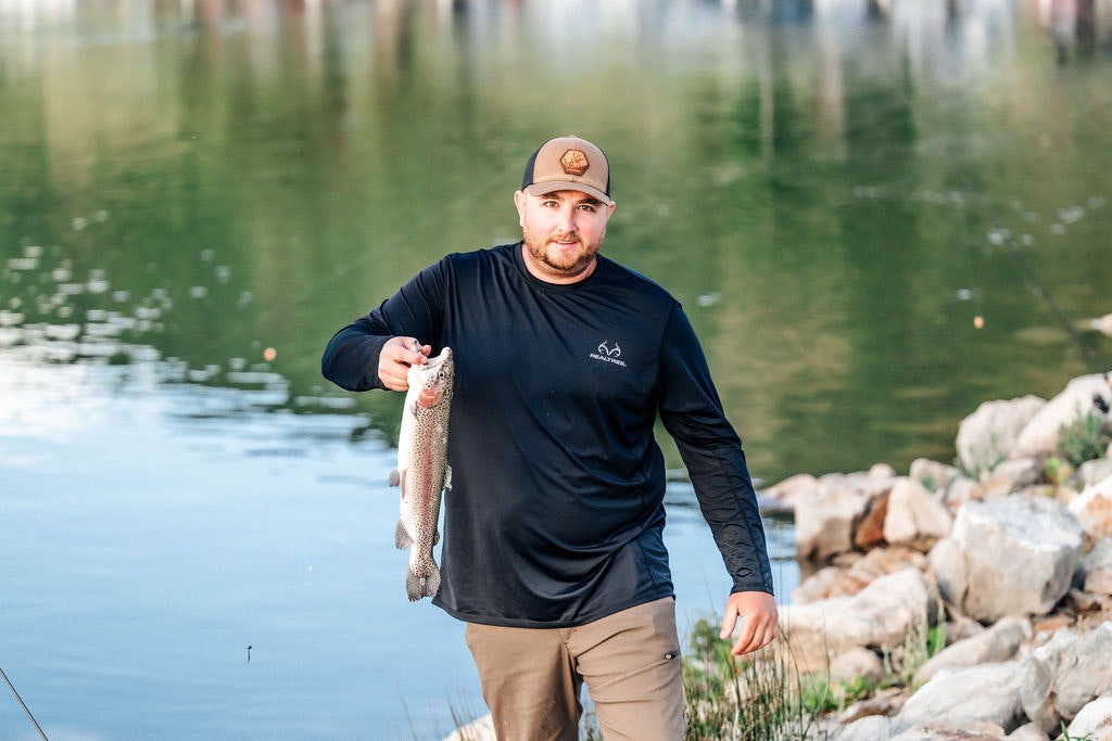 A man showing a fish he caught by a lake, with rocks and reflections on the water.