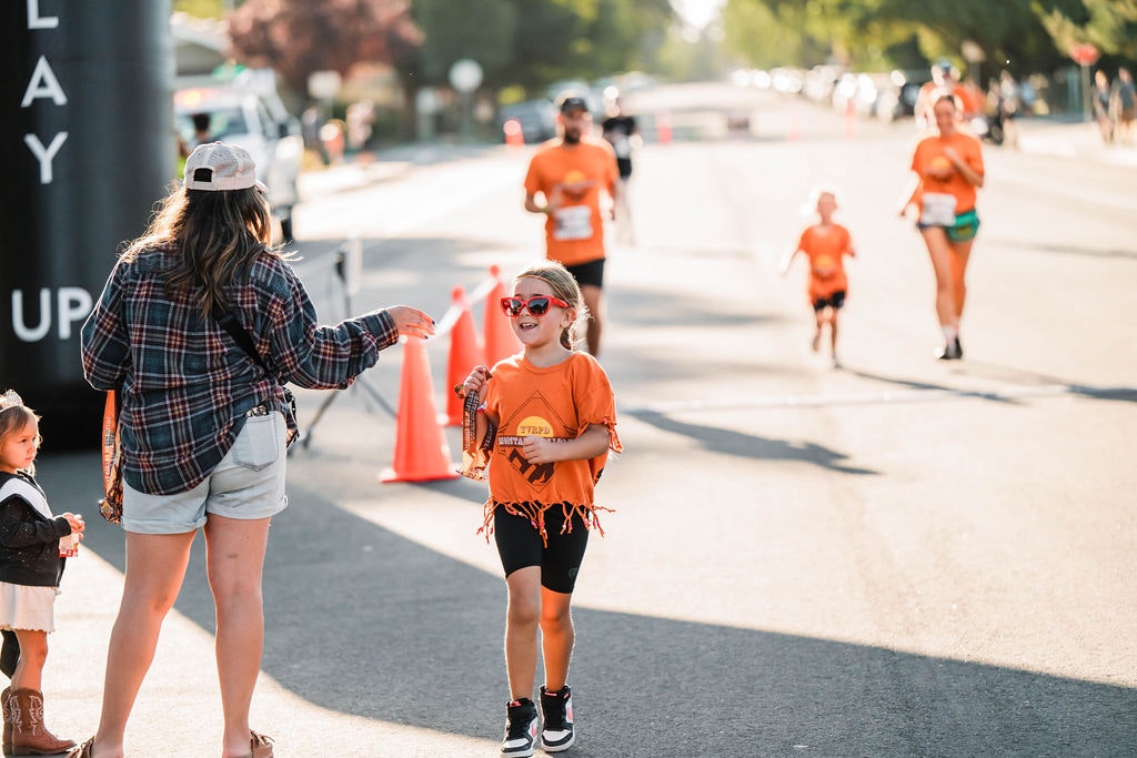 A child in orange, wearing sunglasses, finishes a race, receiving a medal from a woman. Others in orange shirts run in the background.