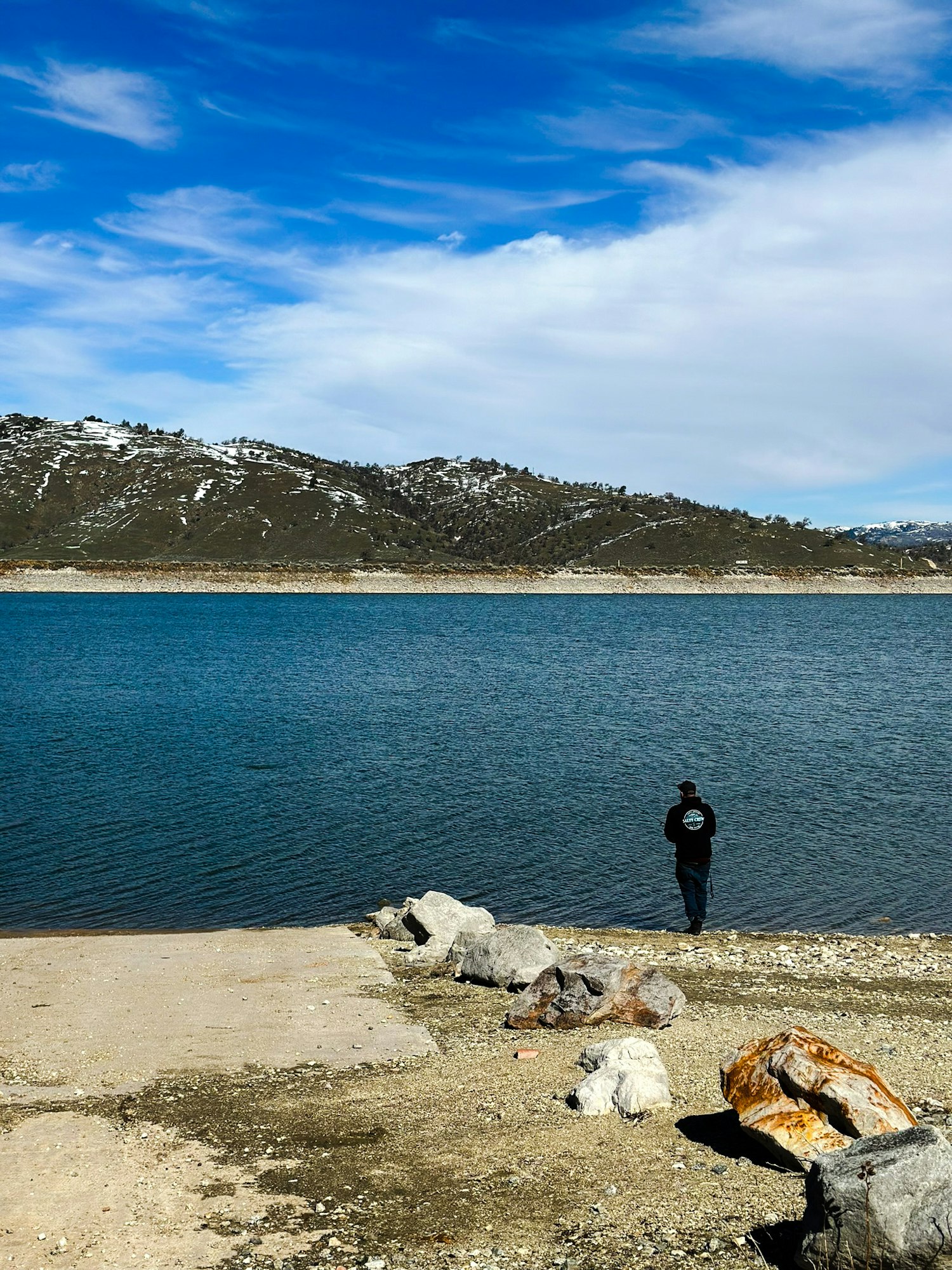 A person stands by a lake with snowy hills in the background under a blue sky with clouds.