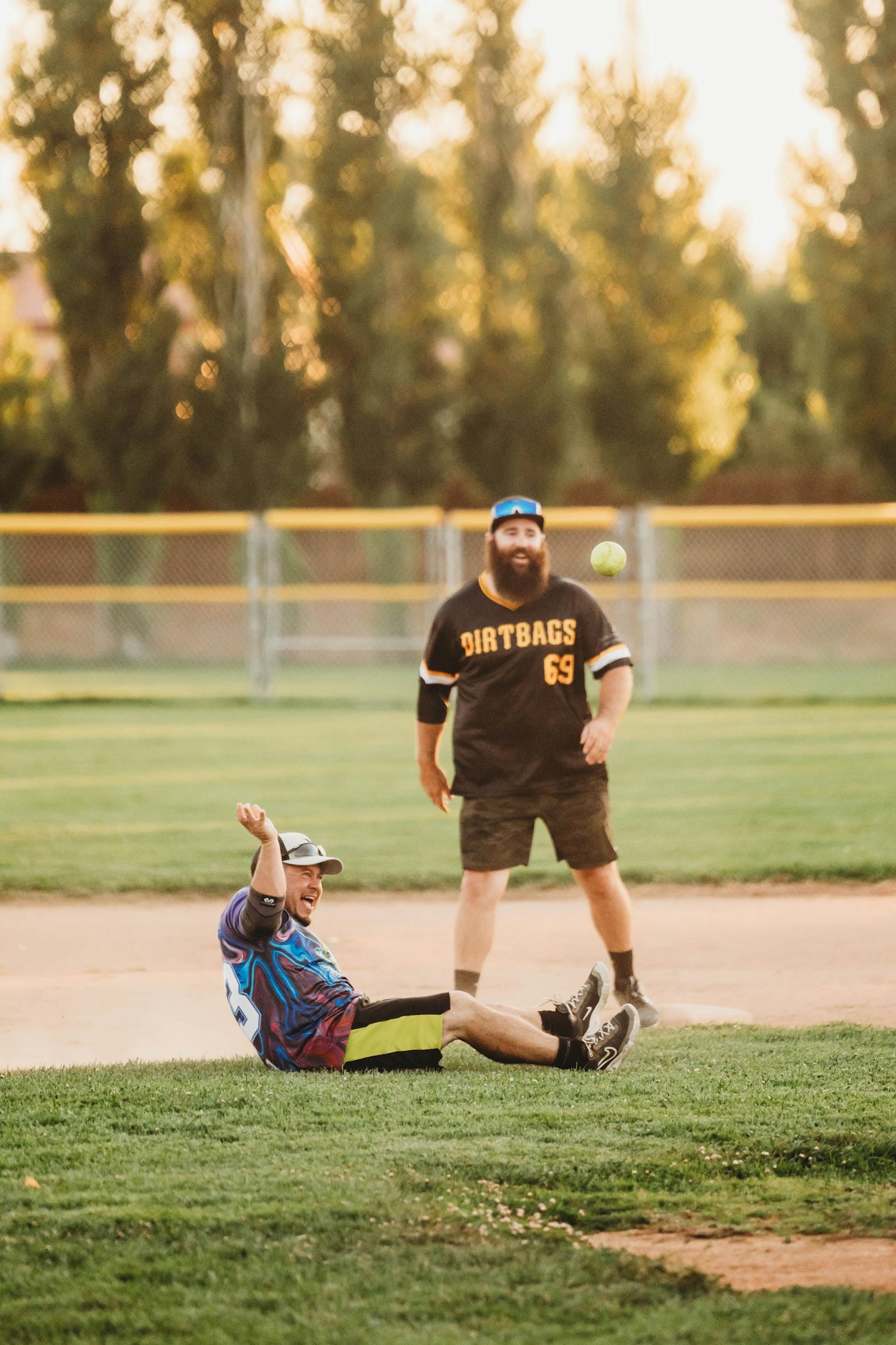 Two people on a baseball field, one sitting on the ground laughing, the other standing with a ball in mid-air.
