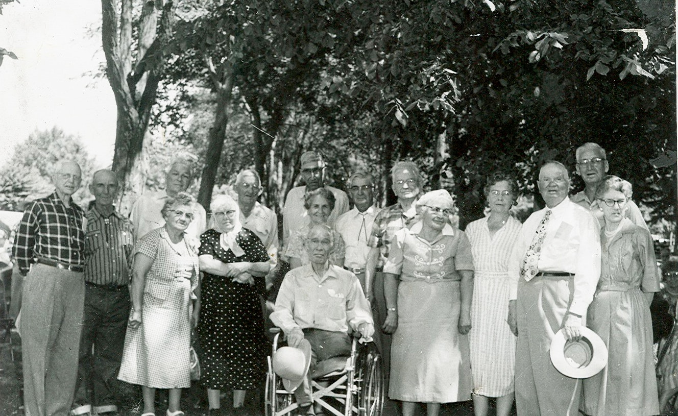 A group of elderly people poses outdoors under trees, and one person is seated in a wheelchair.