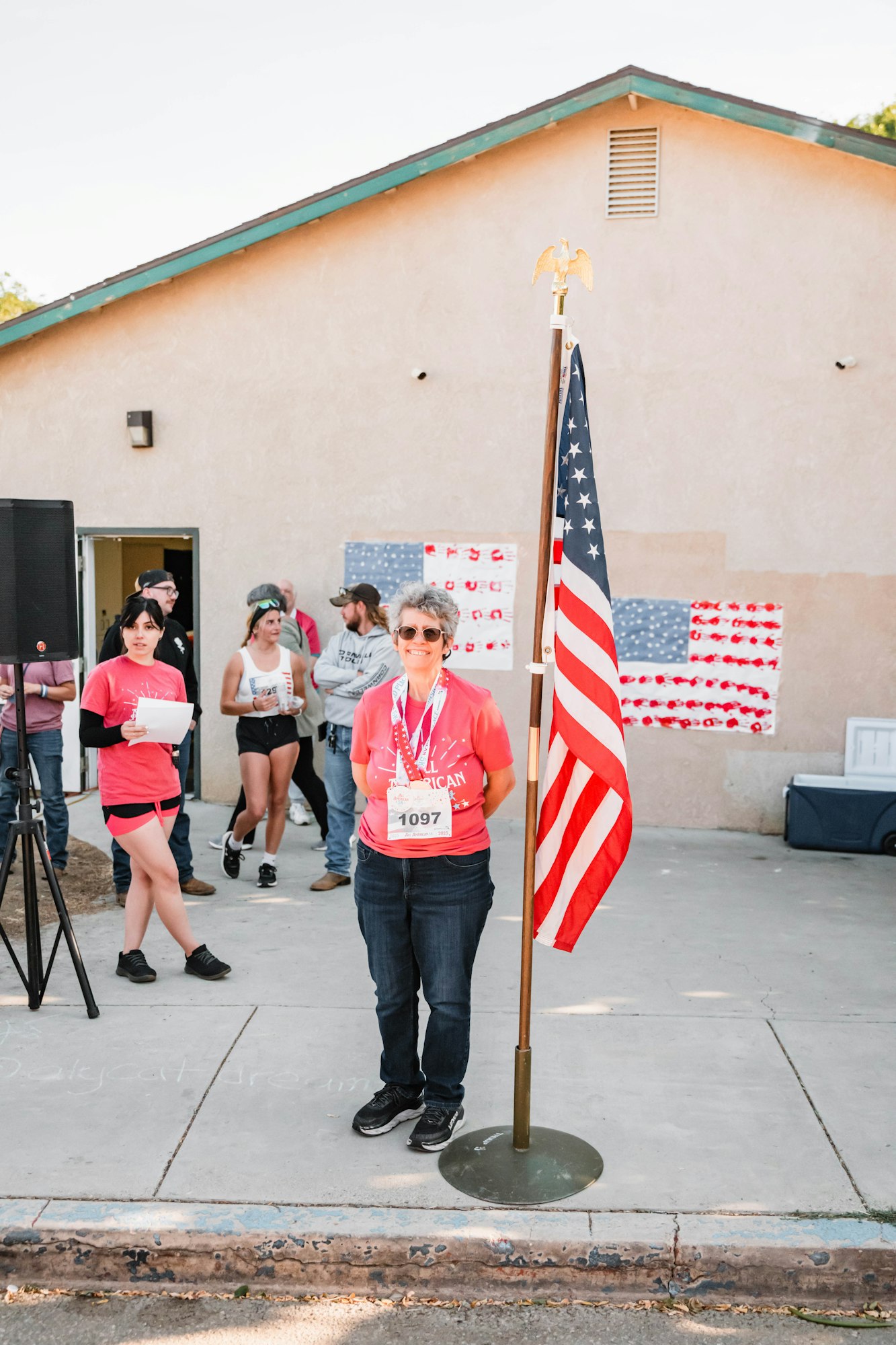 Person with medals stands near a flag, others gather casually nearby; building in background with a flag-themed decoration.