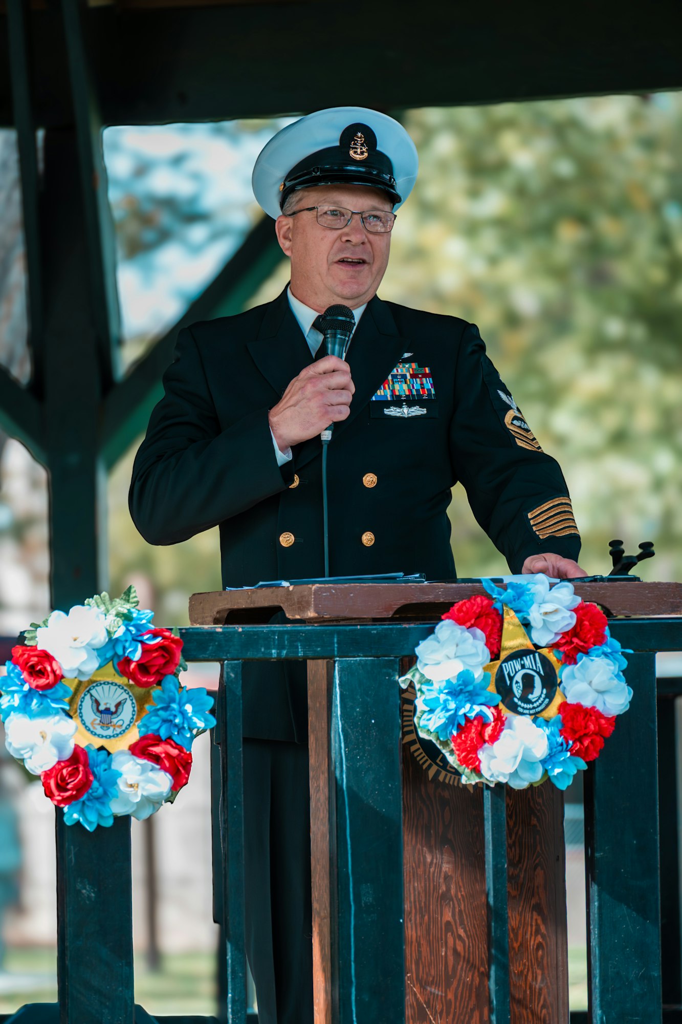 A uniformed person speaks at a podium adorned with floral wreaths.
