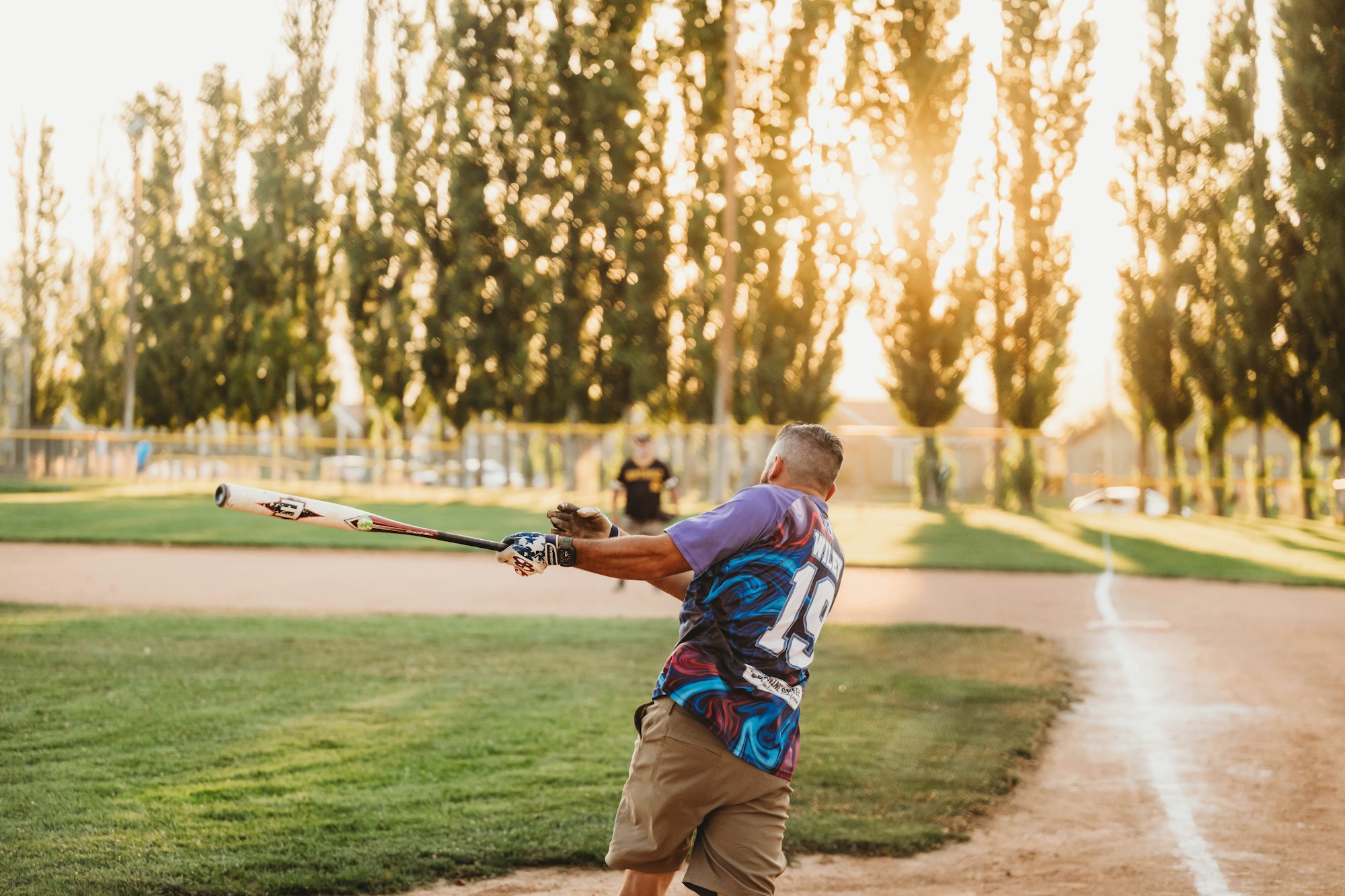 A person swinging a baseball bat at sunset on a dusty field with trees in the background.