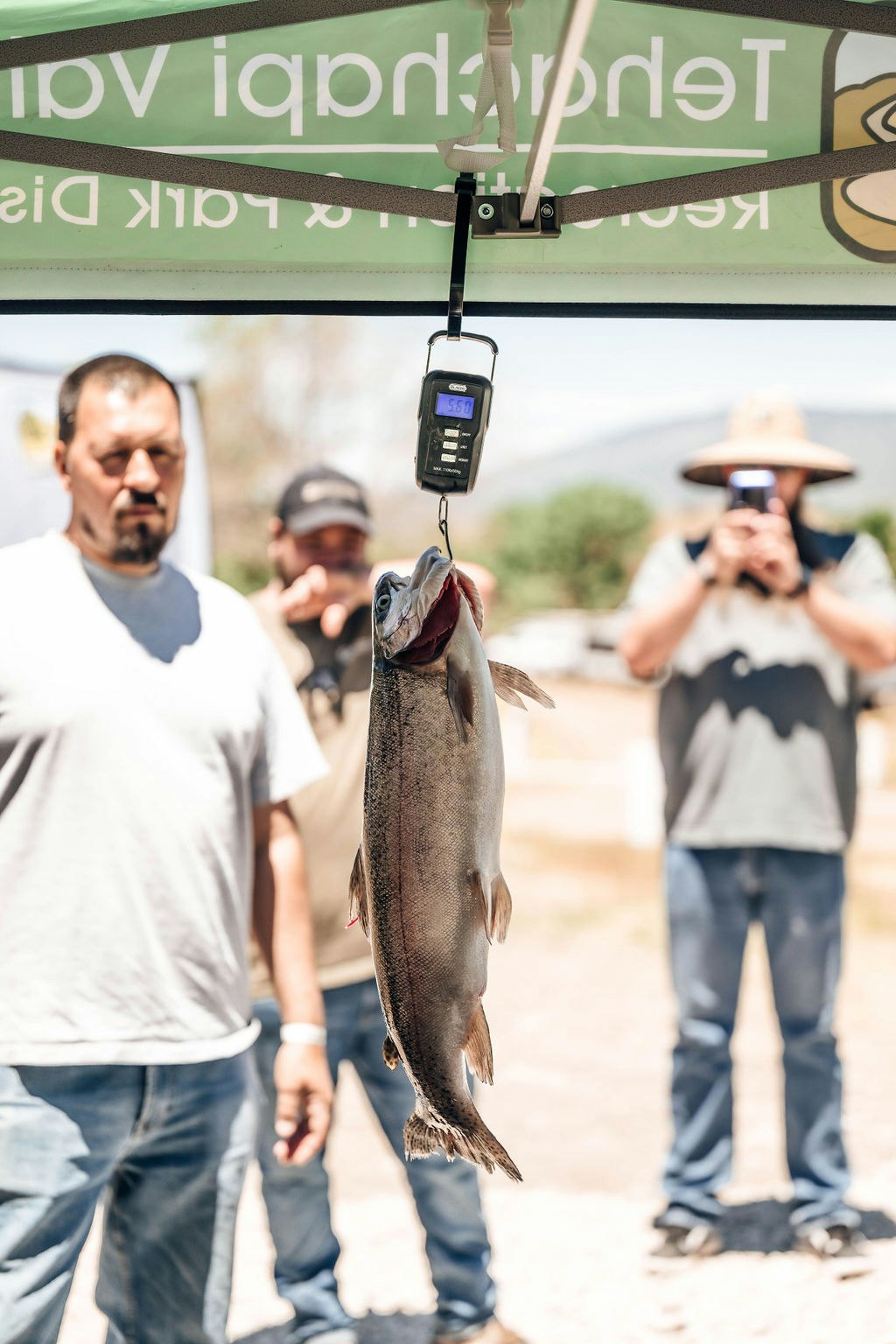 A fish being weighed with a digital scale, with onlookers in the background.