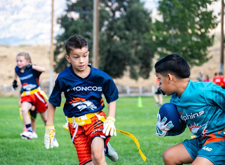 Kids playing flag football on a grassy field wearing team jerseys.