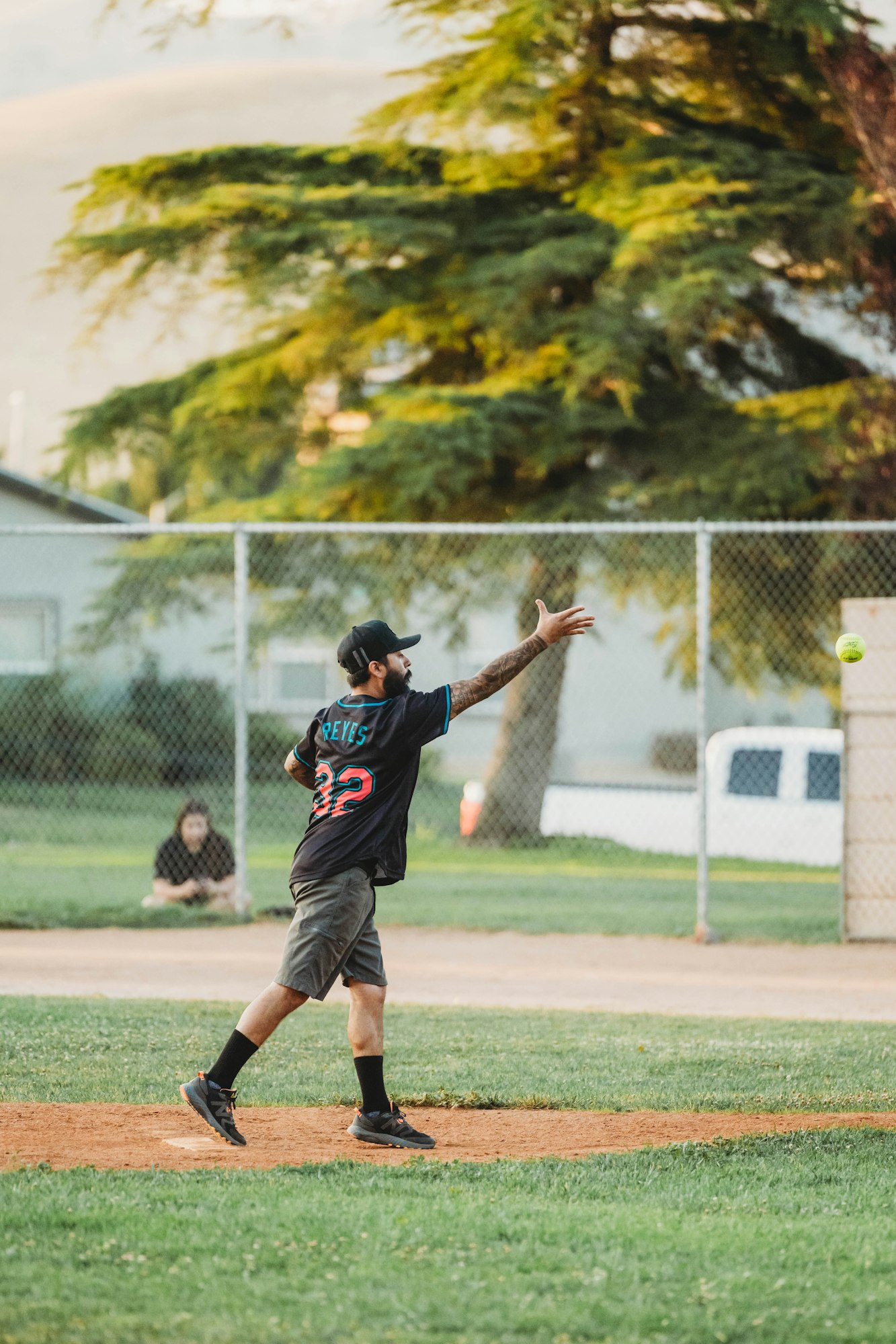 A person is pitching a softball in a park with trees in the background.