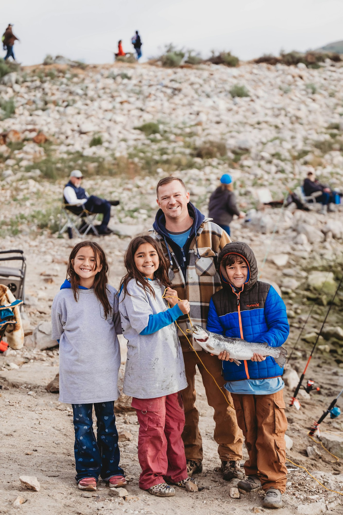 A group of people, including kids holding a fish, are posing happily outdoors on rocky terrain with fishing gear nearby.