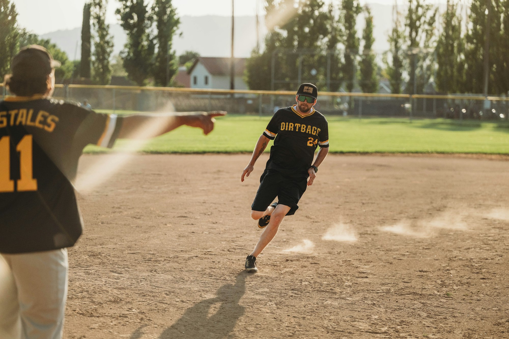 Two baseball players on a dusty field, one running and another pointing, sun setting in the background.
