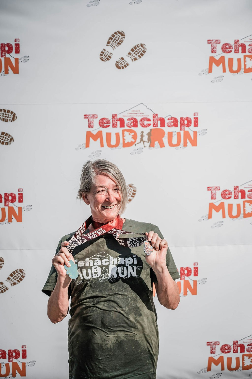 Smiling person holding a medal, wearing a "Tehachapi Mud Run" shirt, in front of a branded backdrop.