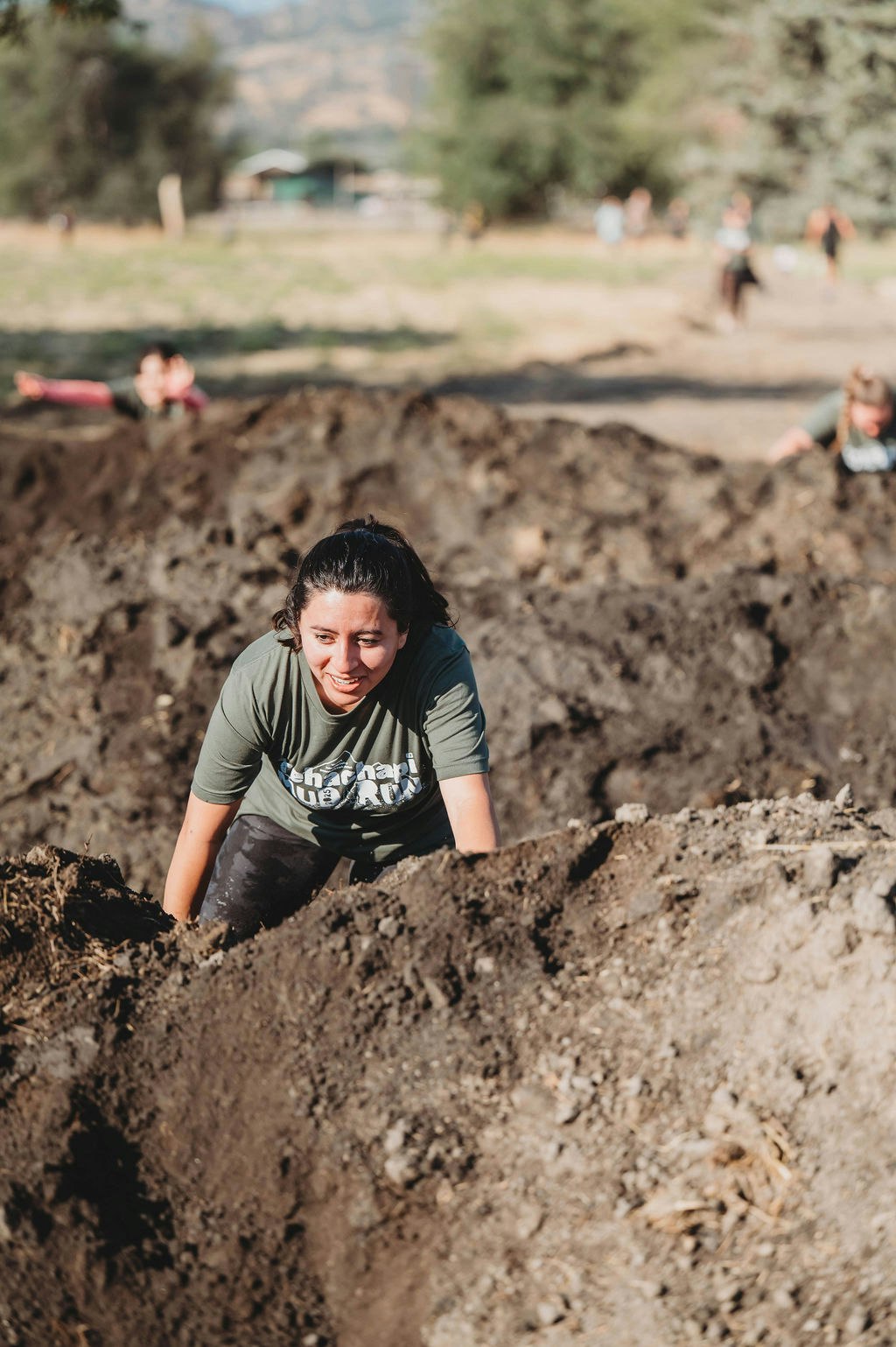 A person in a green shirt participates in a muddy obstacle race, climbing over dirt with others in the background.